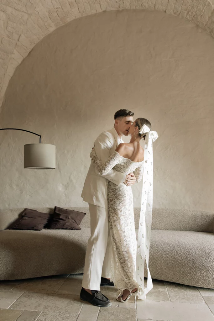 A bride and groom share a tender kiss beneath arched stone walls in an elegant, minimalist Puglia wedding setting.