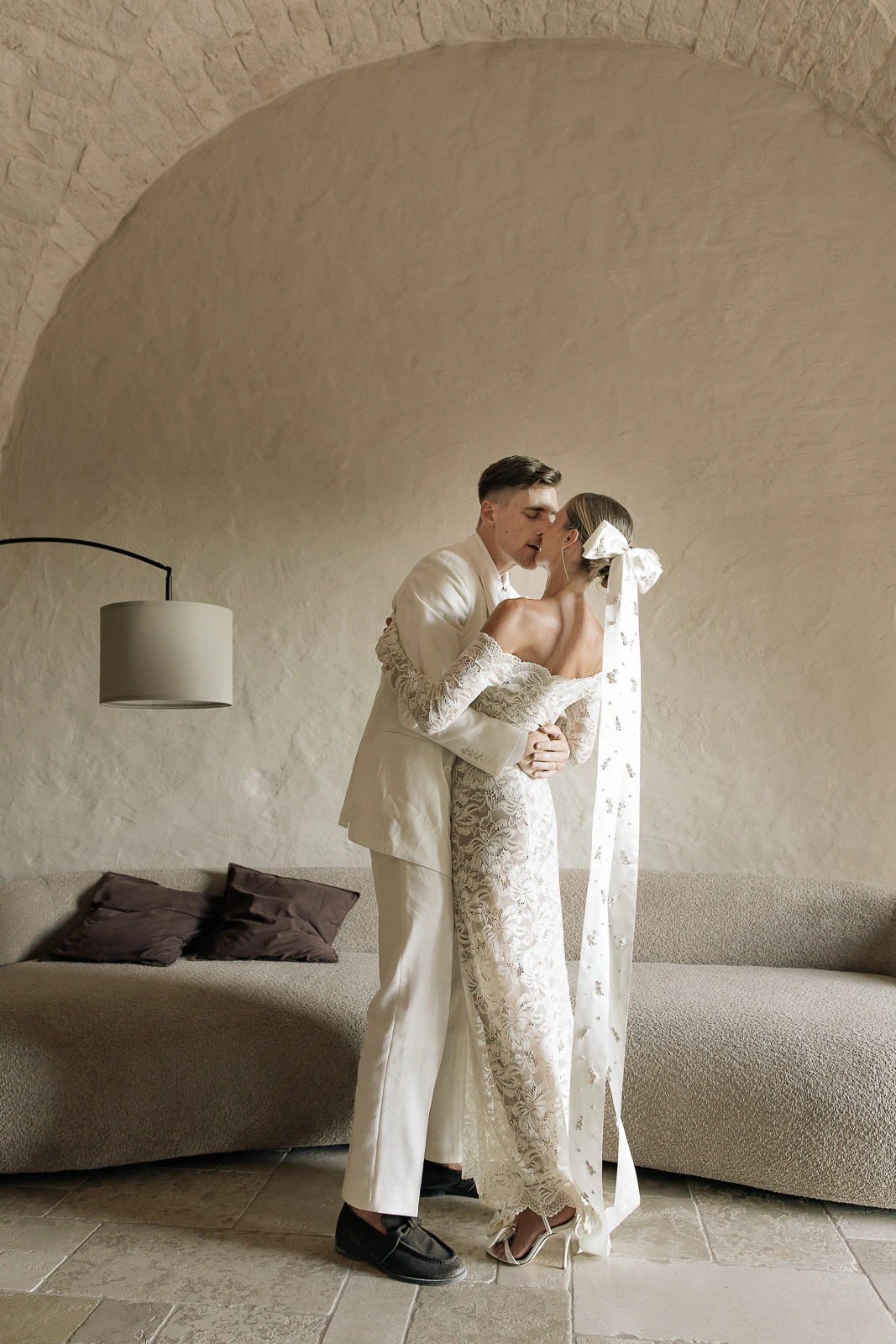 A bride and groom share a tender kiss beneath arched stone walls in an elegant, minimalist Puglia wedding setting.
