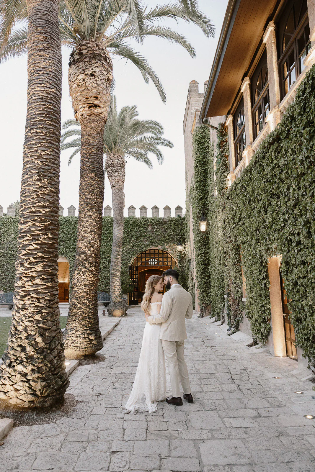 A bride and groom share an intimate embrace beneath palm trees by an ivy-clad villa in sunlit Puglia, Italy.