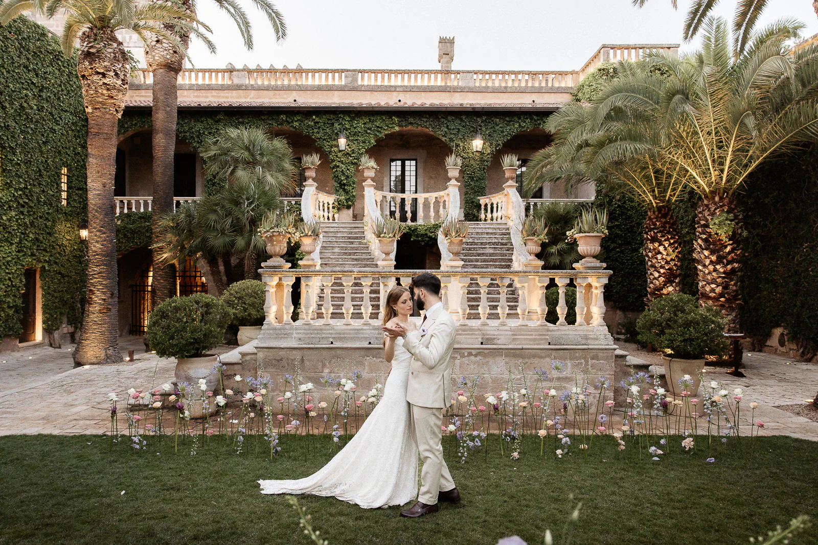 A joyful couple dances on a lush lawn before an ivy-clad Puglia villa, enveloped by blooms in a refined Italian garden.