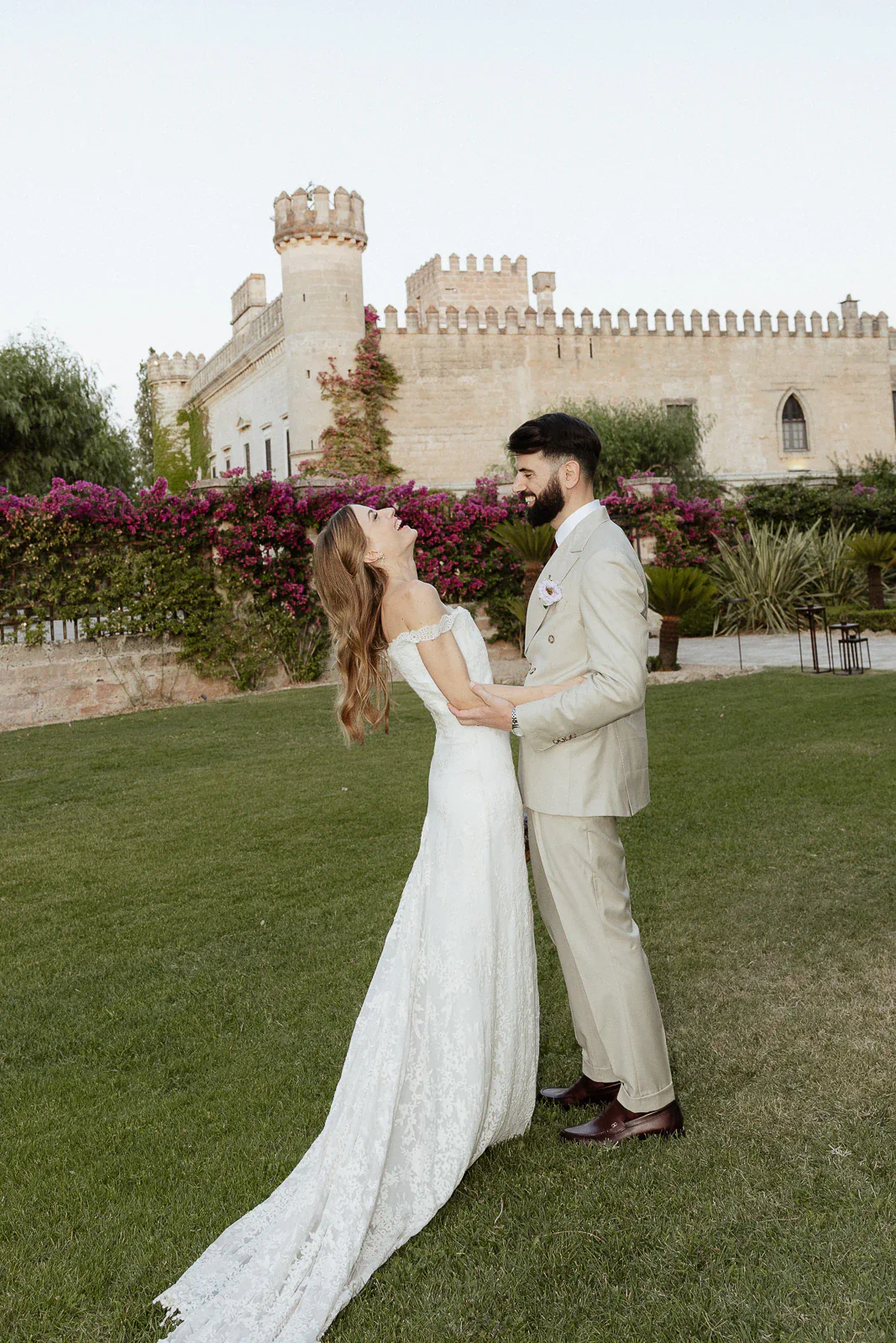 In Puglia, a joyful bride and groom share an intimate moment beside a historic stone villa and lush flowering gardens.