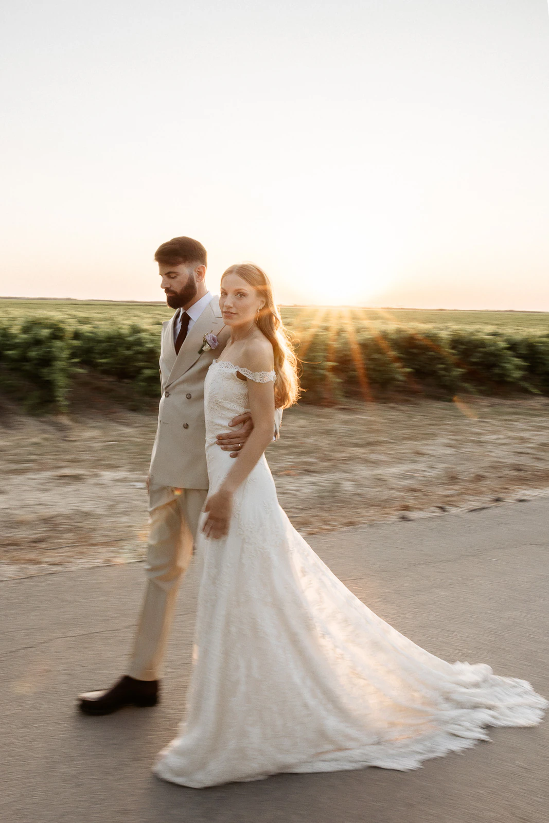 Newlyweds stroll hand in hand along a sunlit Puglian road, their silhouettes framed by golden fields at sunset.