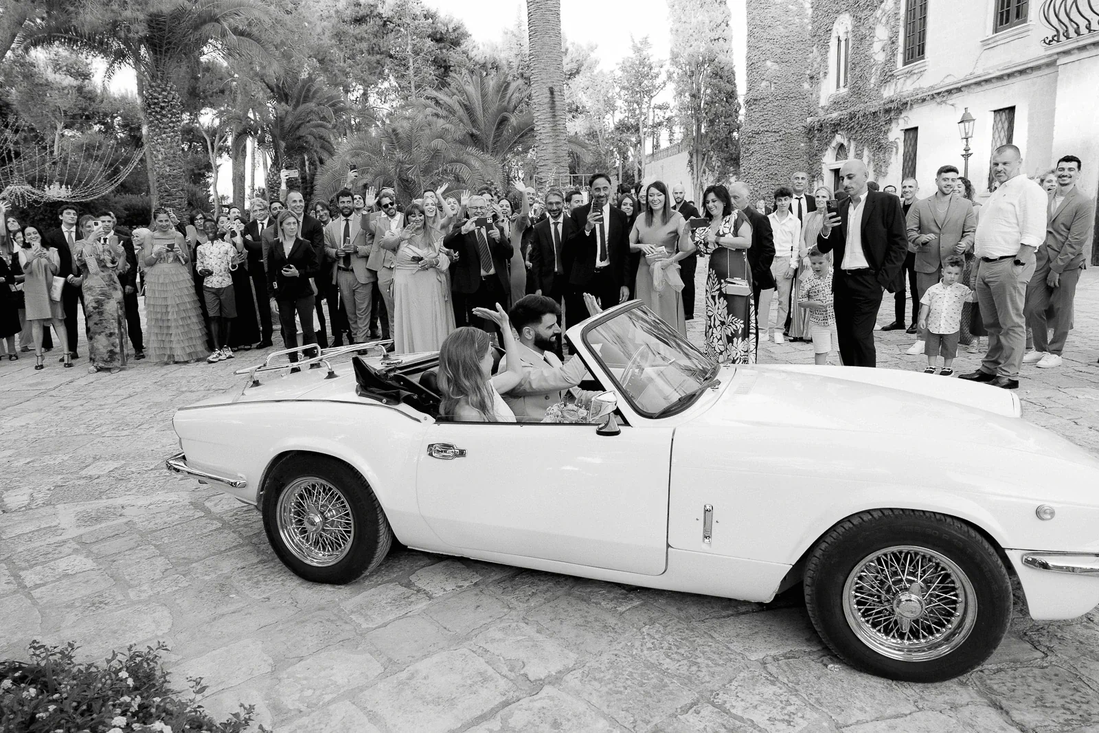 Guests look on as a couple shares an intimate moment in a white convertible, framed by Italian architecture and cobblestone charm.