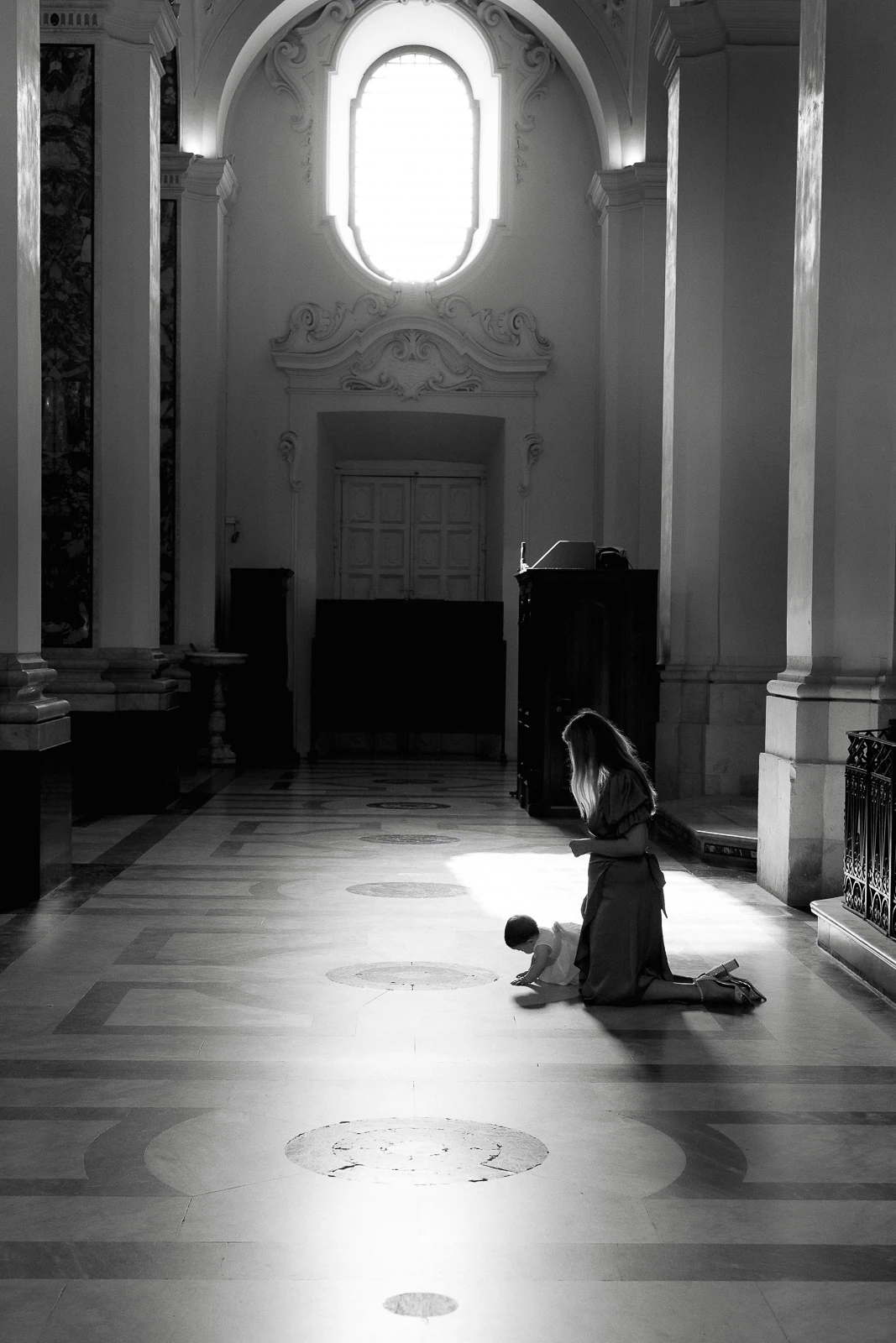 In a sun-drenched Puglia church, a woman lovingly kneels beside a child, framed by soaring ceilings and arched windows.