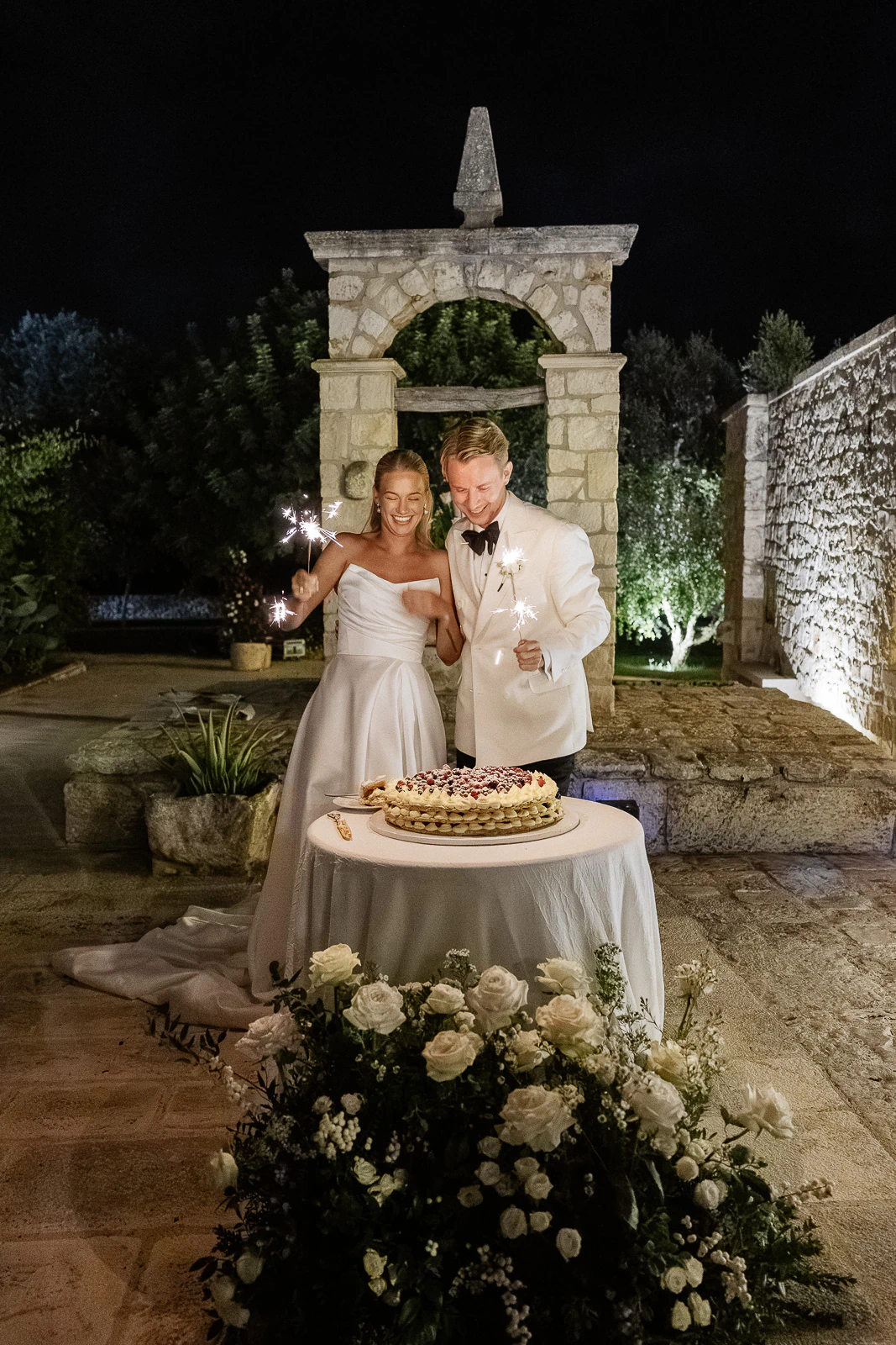 A joyful bride and groom celebrate under Puglian night skies, holding sparklers beside their elegantly adorned wedding cake.