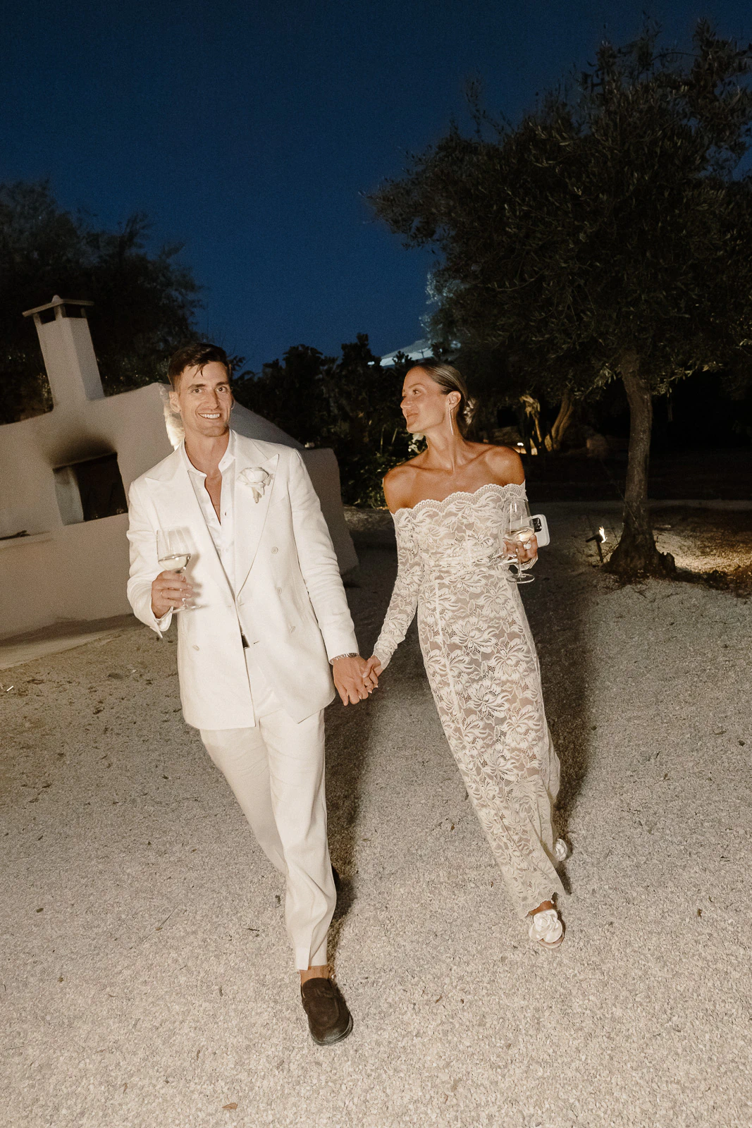 An elegant couple in white strolls hand-in-hand along a starlit gravel path in Puglia, Italy, savoring the evening.