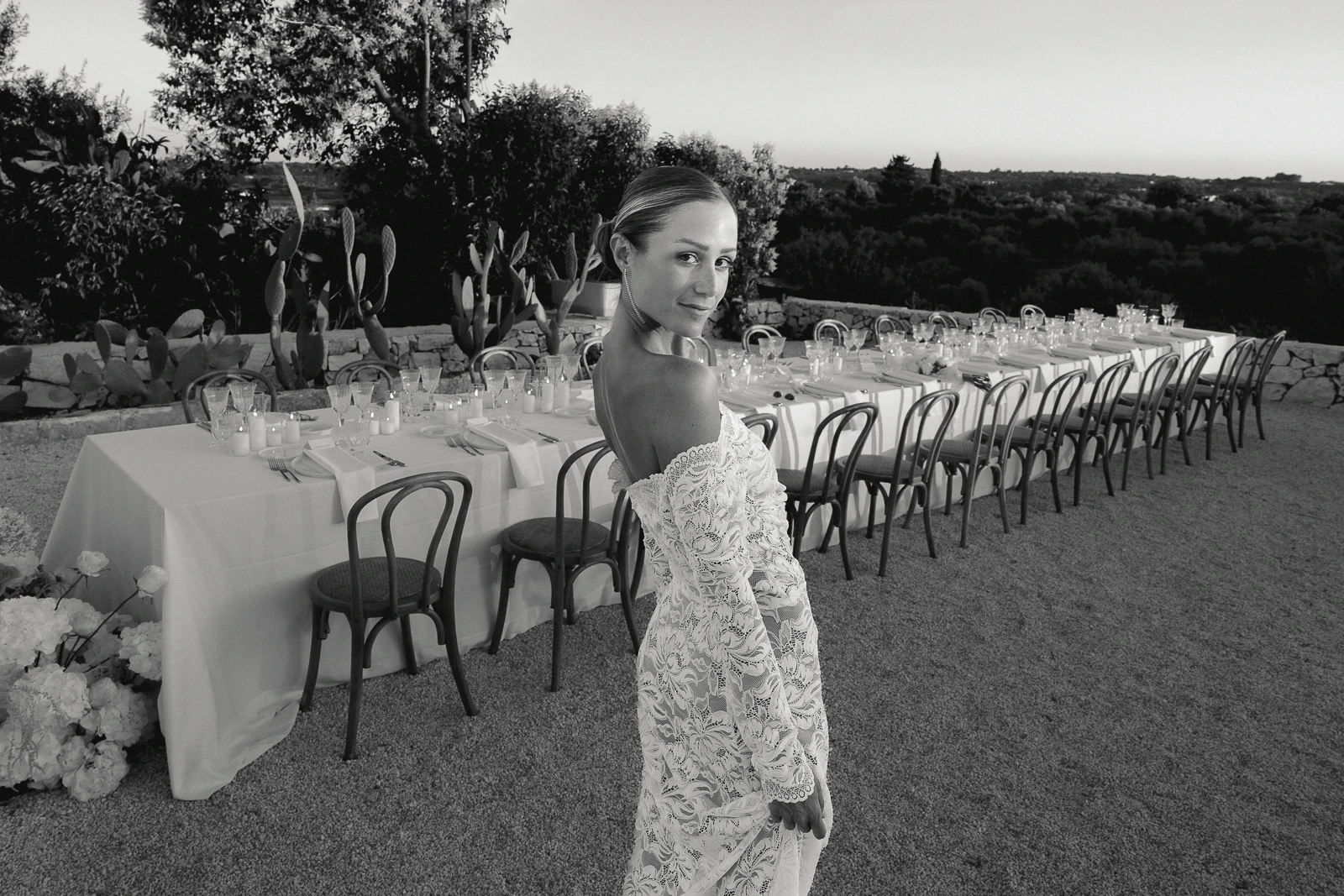Amid Puglia’s sunlit landscape, a bride in elegant lace stands by an alfresco reception table, ready to welcome loved ones.