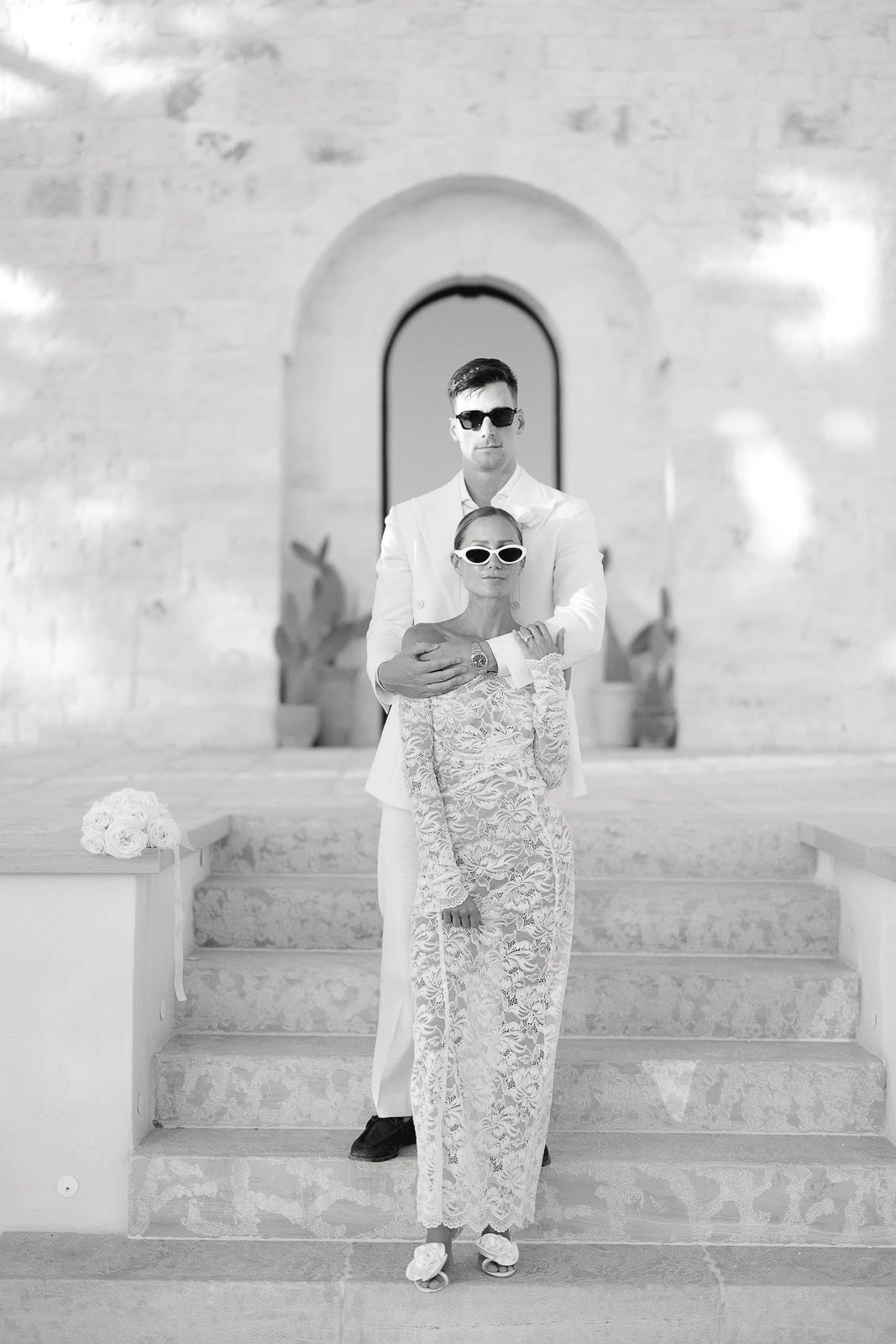 An elegant couple in sunglasses pose on sunlit stone steps in Puglia, radiating effortless connection and Italian sophistication.
