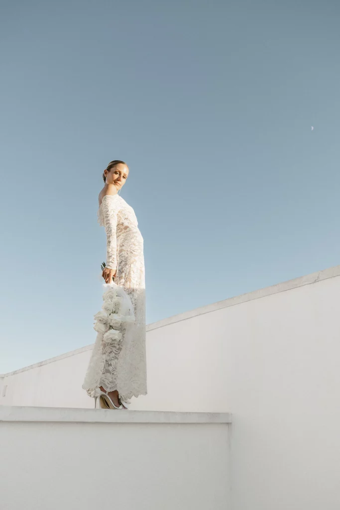 A bride in a lace gown stands on sunlit white stone in Puglia, Italy, bouquet in hand beneath a radiant blue sky.