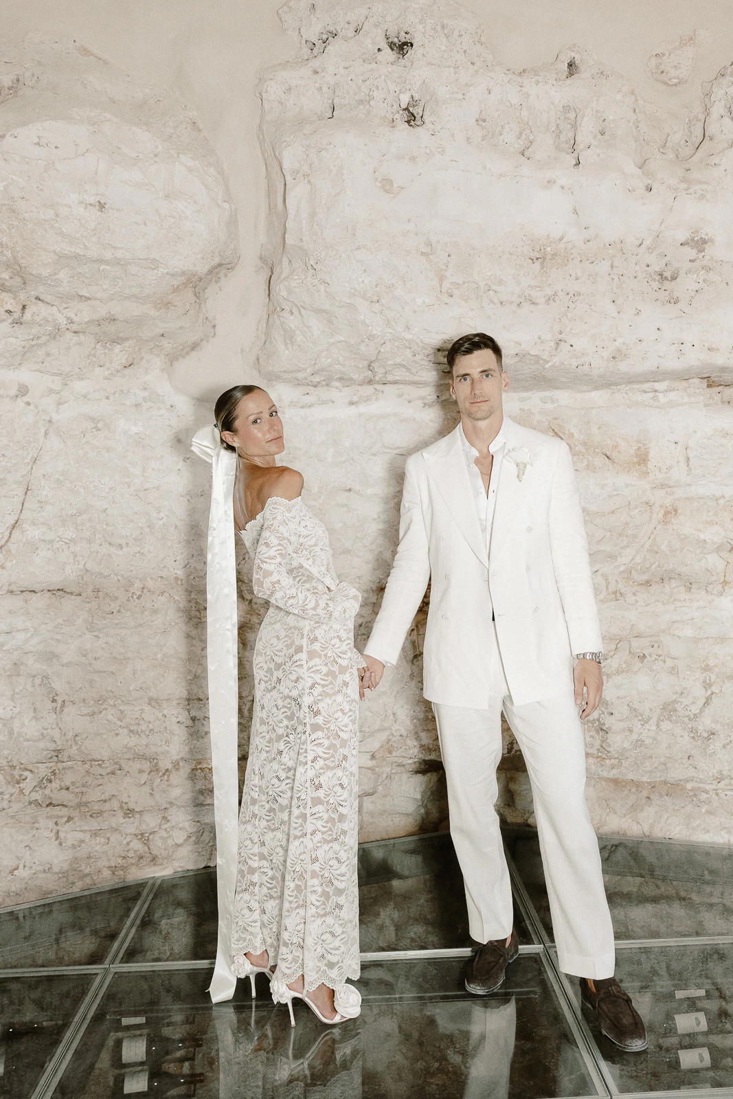 An elegant couple holds hands before a timeless stone wall in Puglia, her lace gown and ribbon evoking effortless Italian romance.