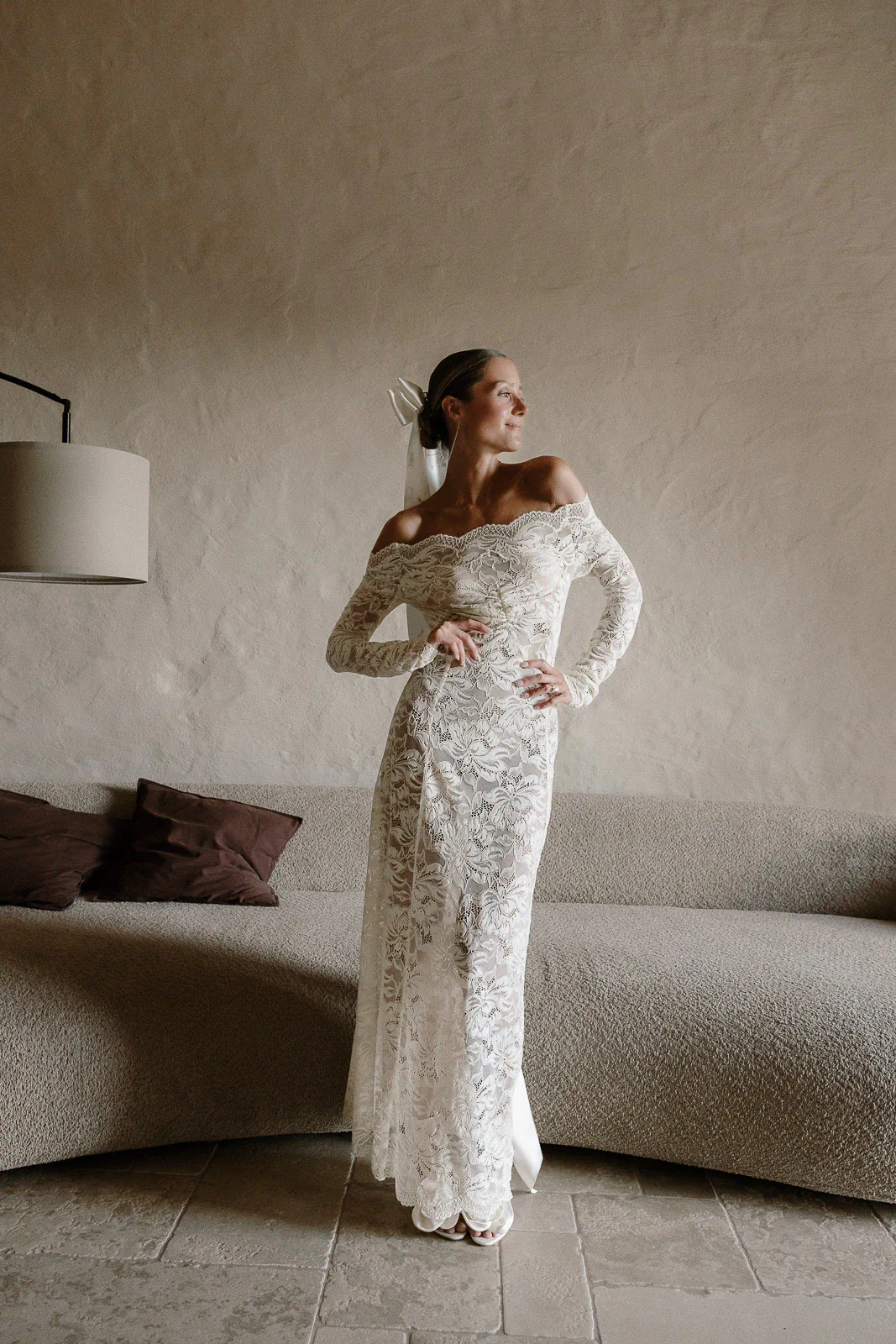 A bride in an off-the-shoulder lace gown stands poised in a serene, minimalist Puglia suite, elegance radiating effortlessly.
