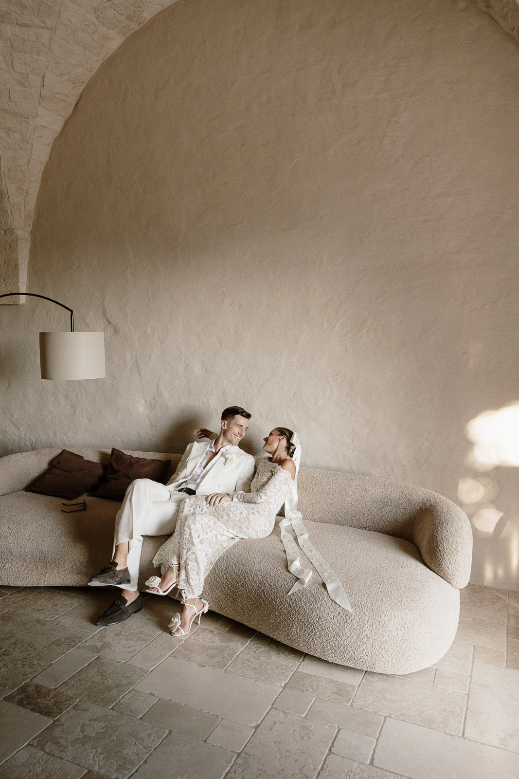 An elegant couple shares an intimate moment on a curved sofa in a minimalist Puglia suite, bathed in soft Italian light.
