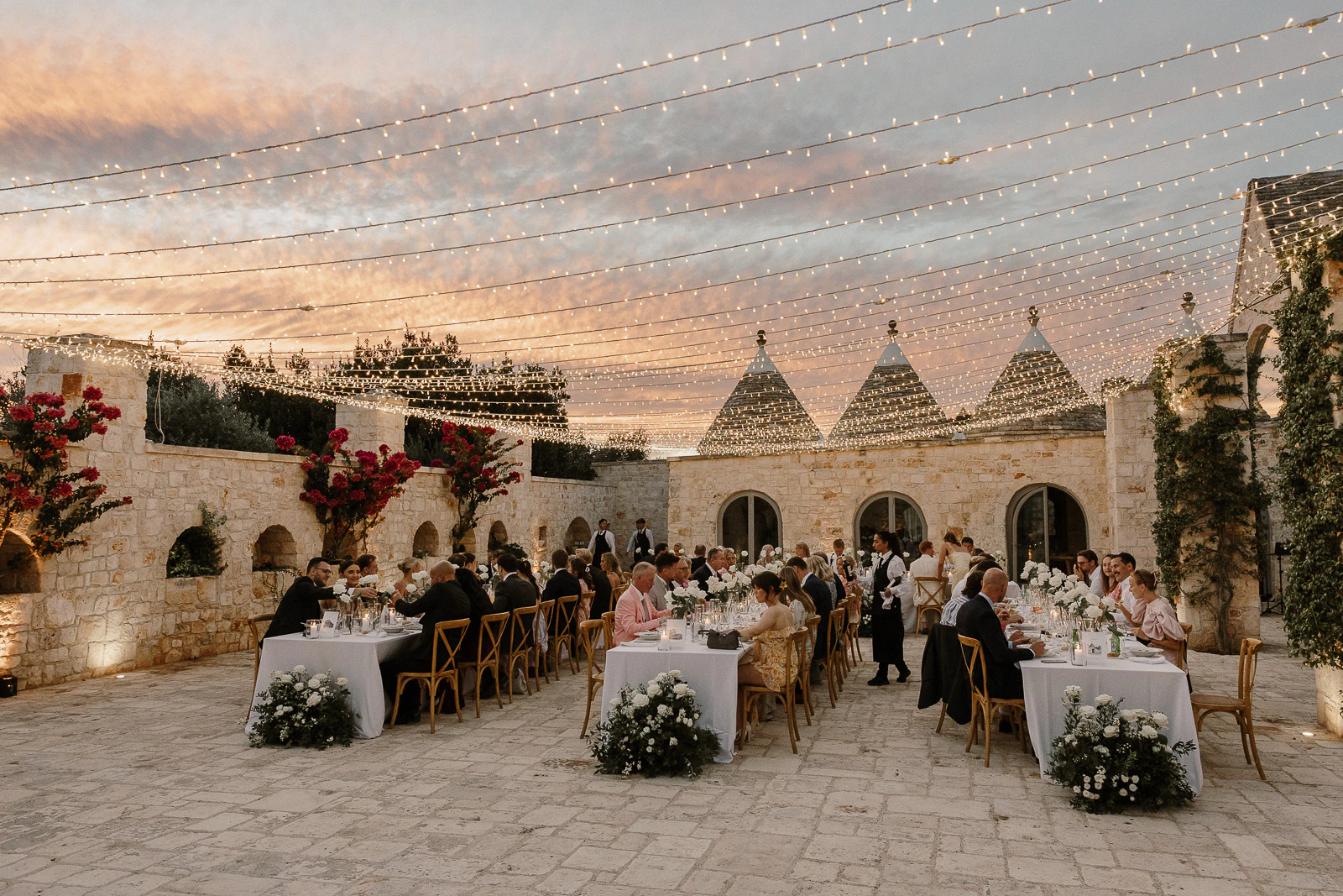 Guests gather in a romantic Puglia courtyard, savoring dinner beneath string lights and sunset skies among iconic trulli rooftops.