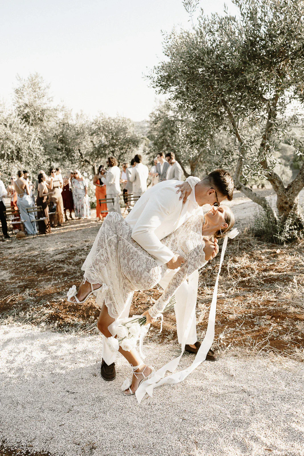 In Puglia, a bride and groom share a passionate kiss beneath olive trees, their loved ones celebrating in sun-dappled elegance.