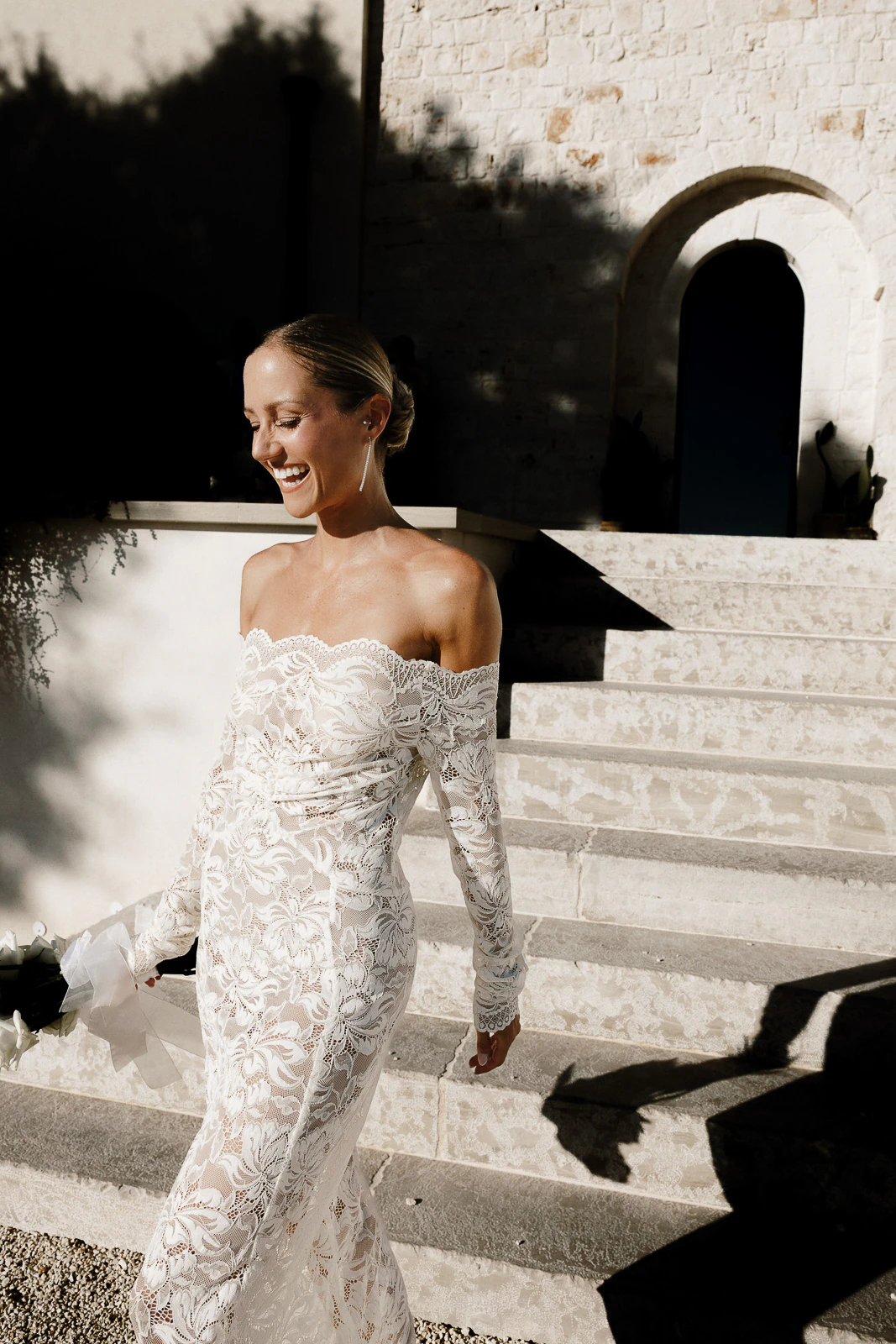A bride in an off-the-shoulder lace gown radiates joy, strolling past sunlit stone steps in picturesque Puglia, Italy.