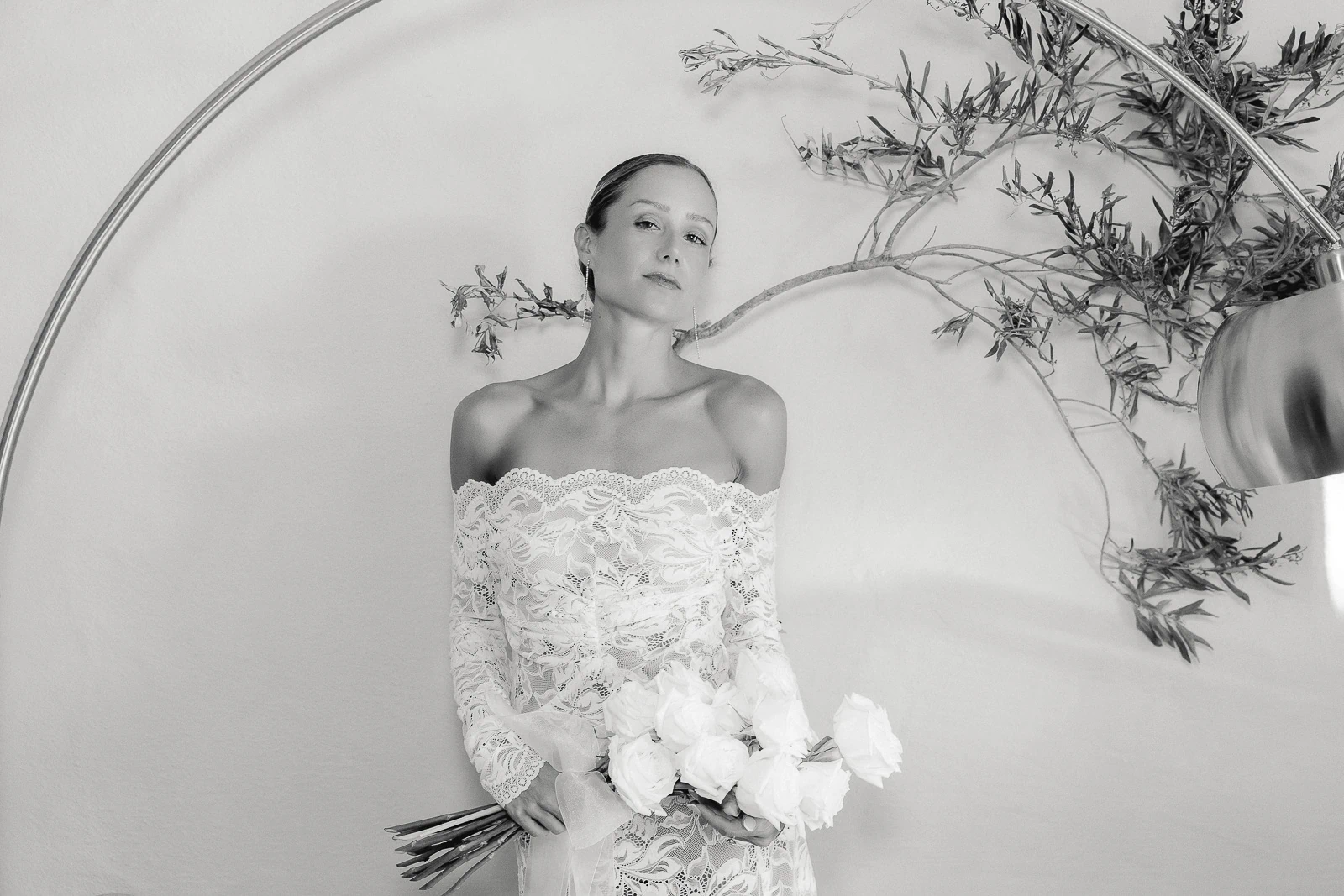 A bride in an elegant lace off-the-shoulder gown cradles white roses, glowing against a serene Italian villa backdrop.