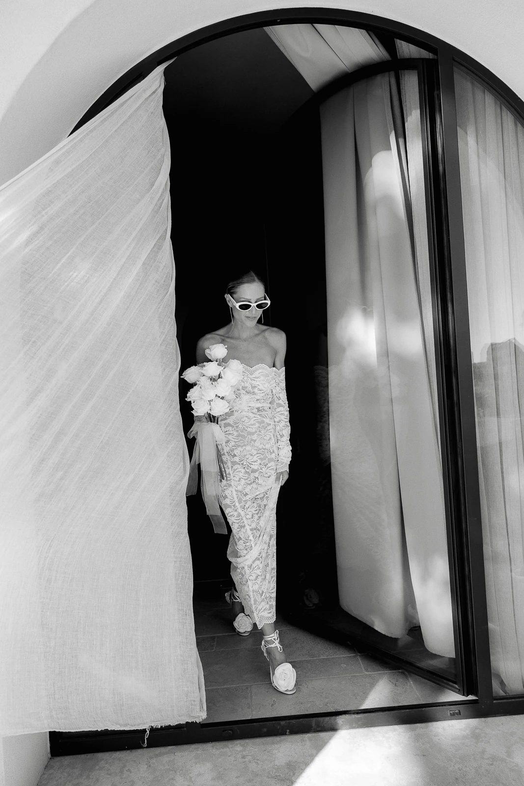 In Puglia, a bride in lace and sunglasses stands in a sunlit doorway, sheer curtains drifting, bouquet in hand.