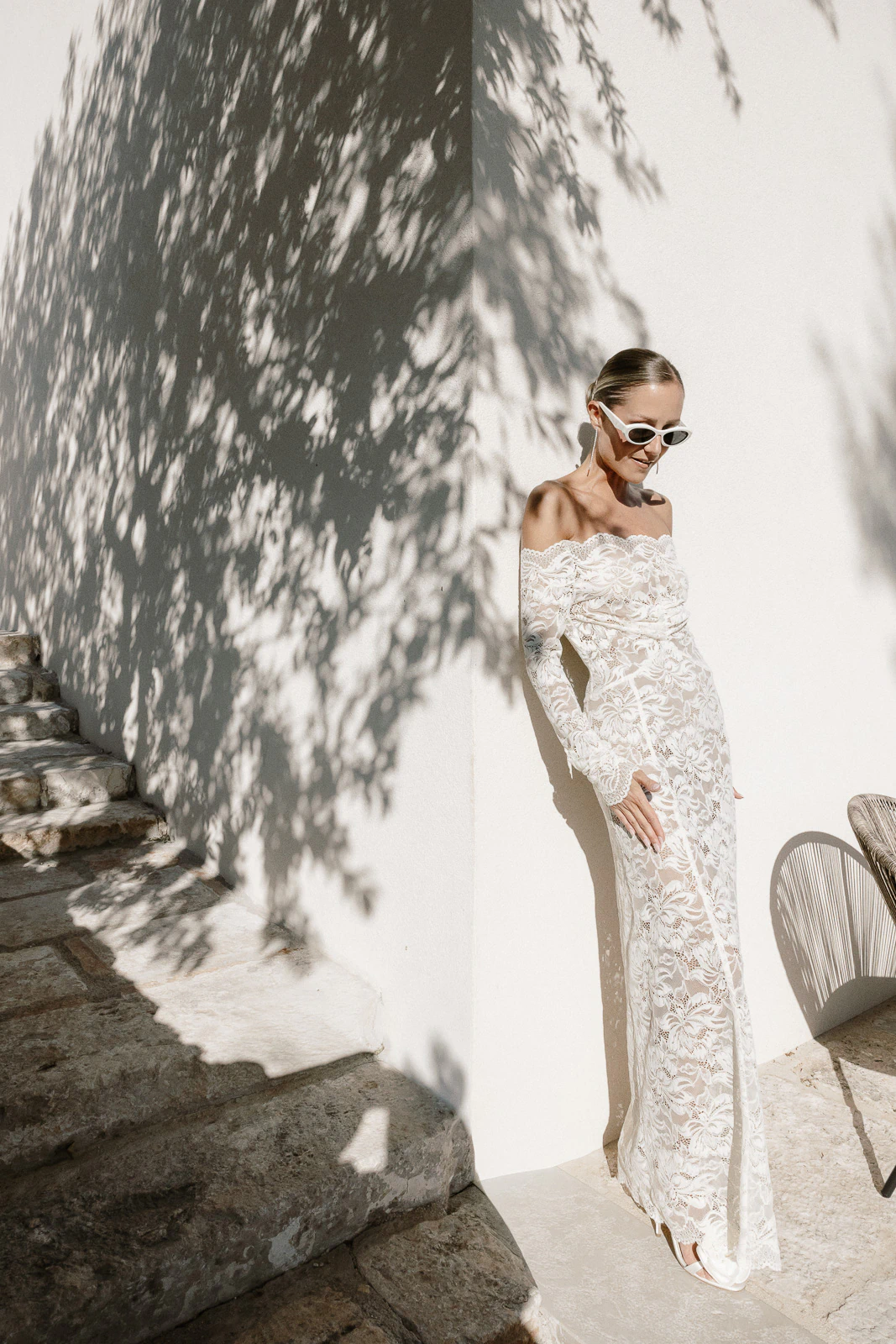 A bride in a refined white lace gown basks in Puglia’s sunlight, framed by leaf shadows on an ancient Italian wall.