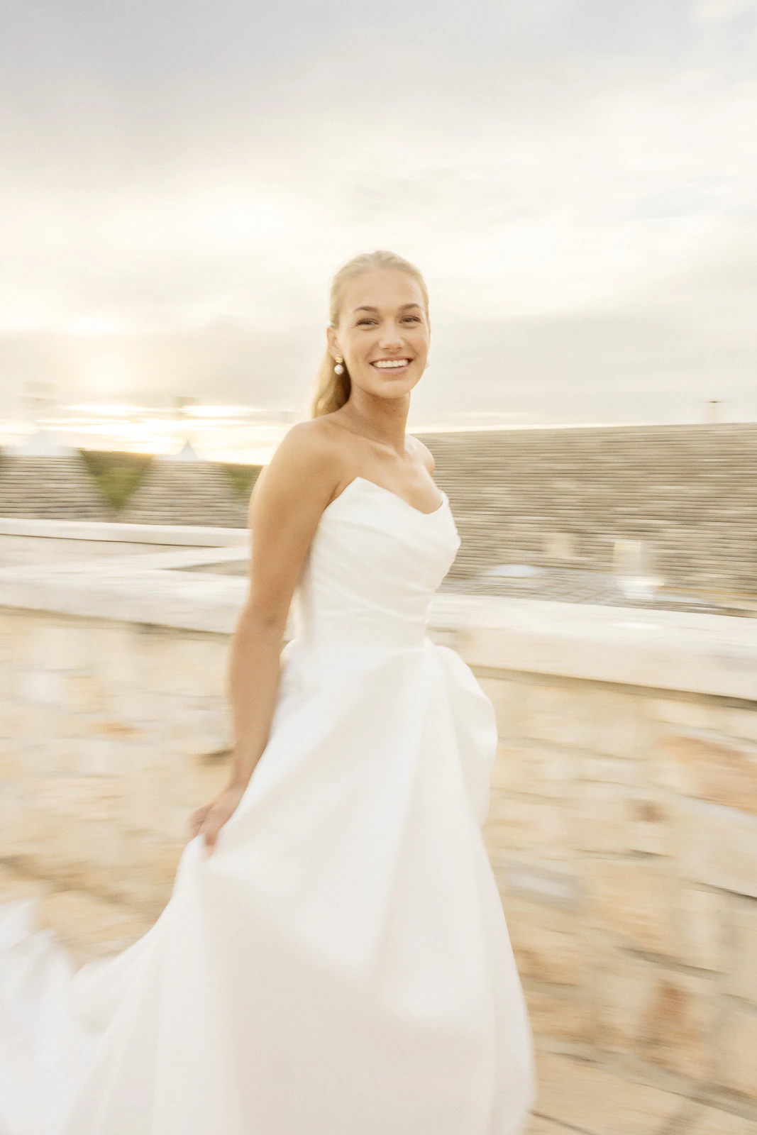 A bride glows in an elegant white gown, framed by rustic stone walls in sunlit Puglia, Italy. Authentic joy radiates.