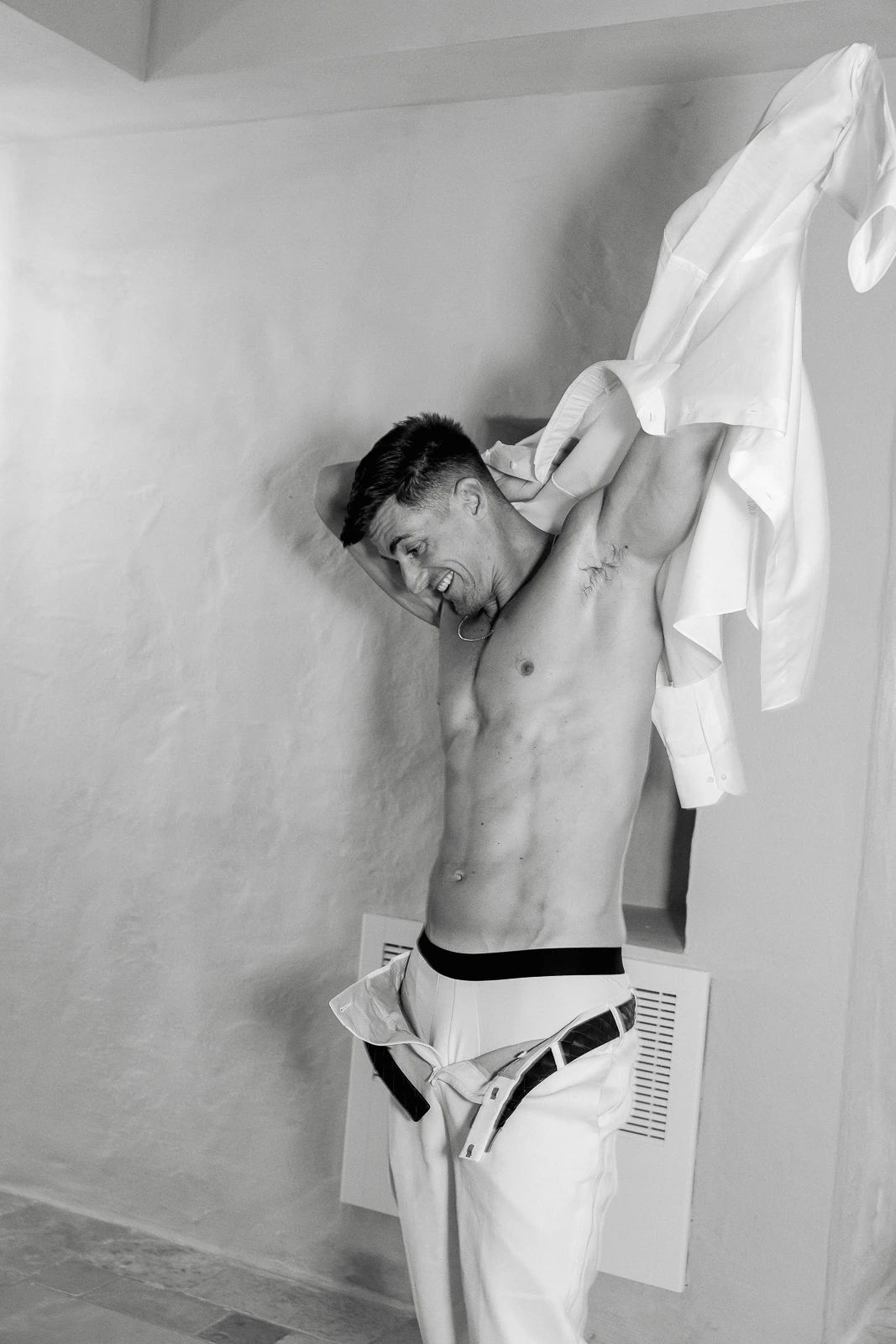 In sunlit Puglia, a groom beams, buttoning his crisp white shirt, anticipation in the air before his elegant Italian wedding.