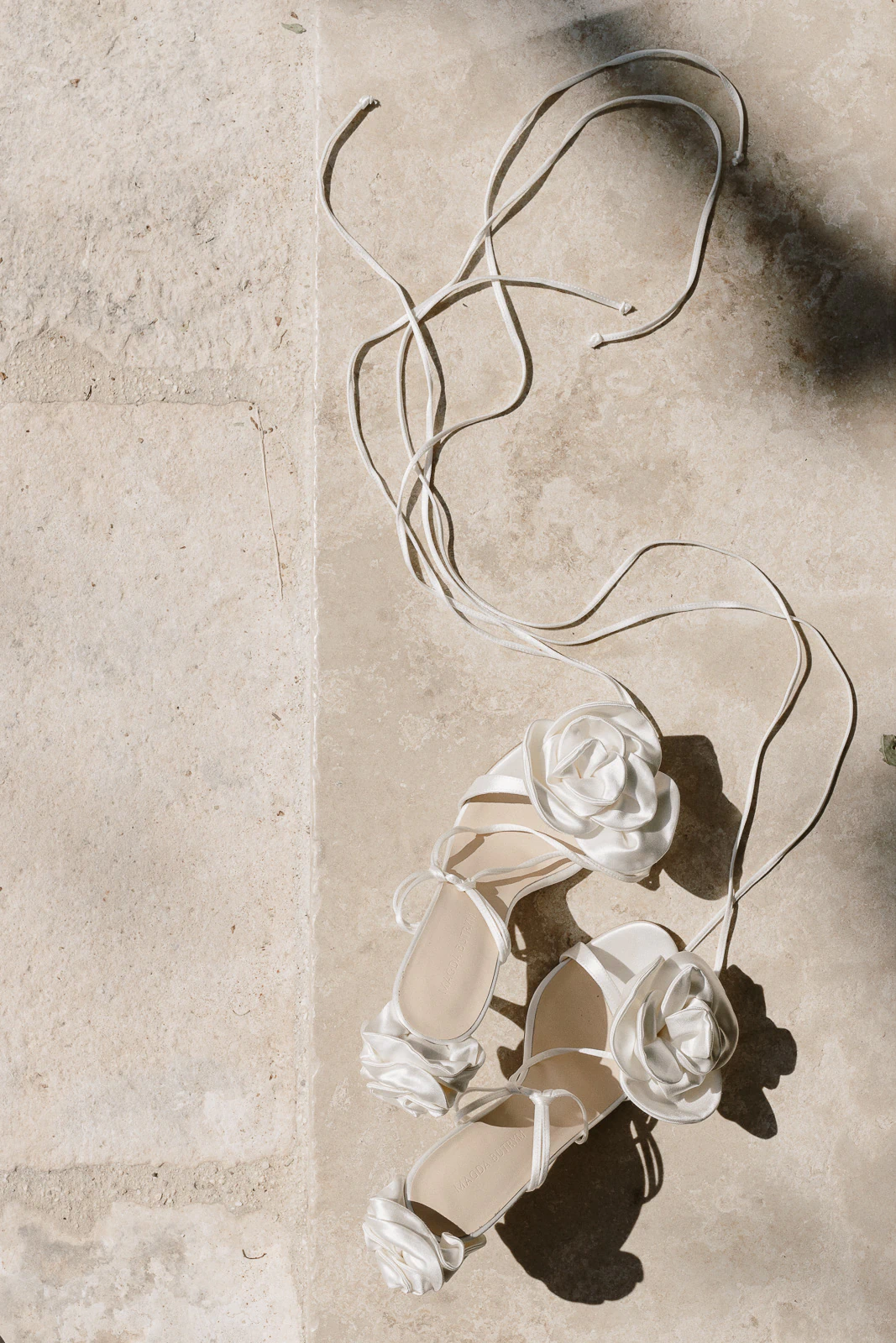 Elegant white floral bridal sandals with delicate lace-up straps, set against sun-warmed Puglia stone—romance meets Italian style.