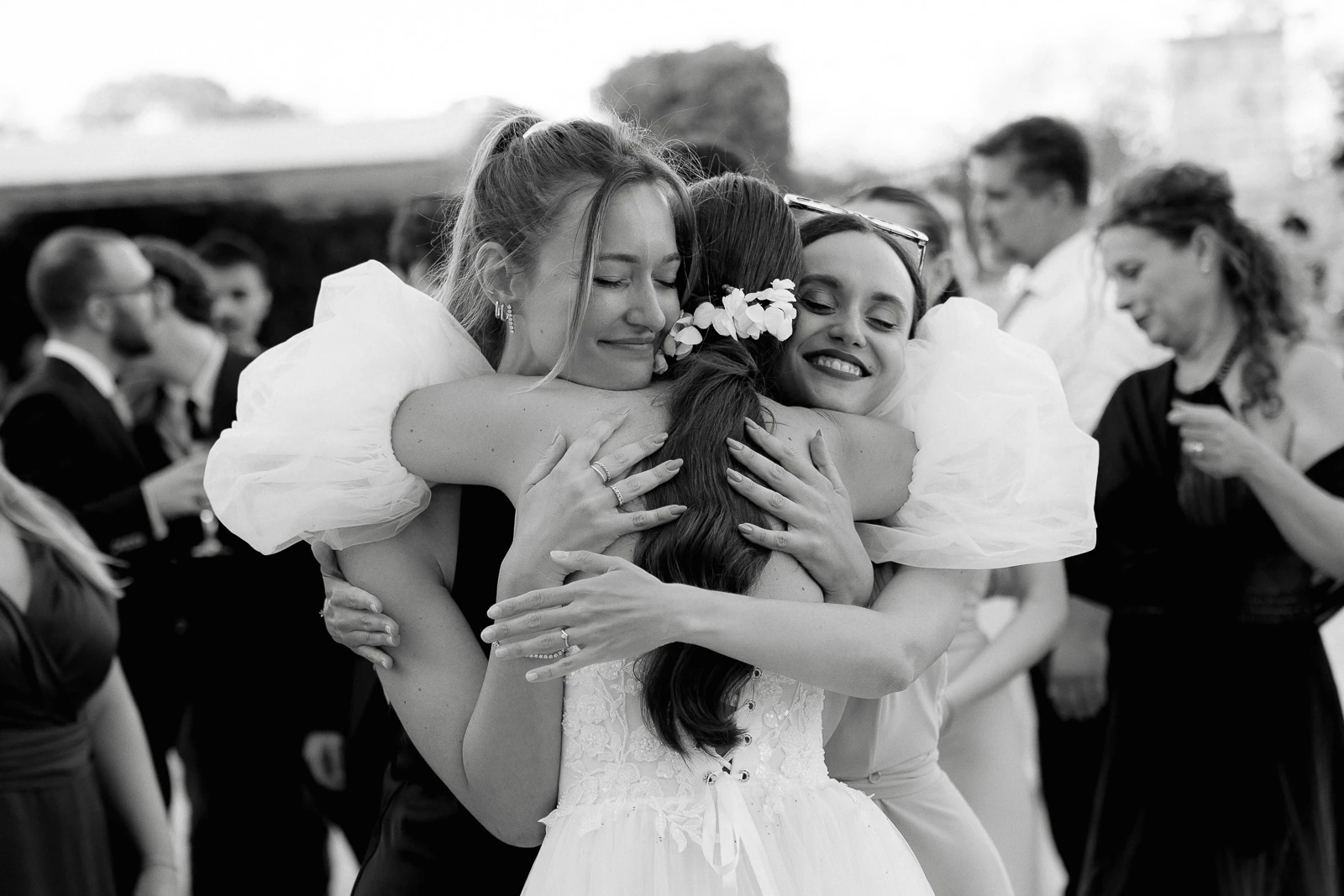 Elegant in formal attire, three women embrace beneath the Puglian sun, their laughter echoing through an intimate Italian celebration.