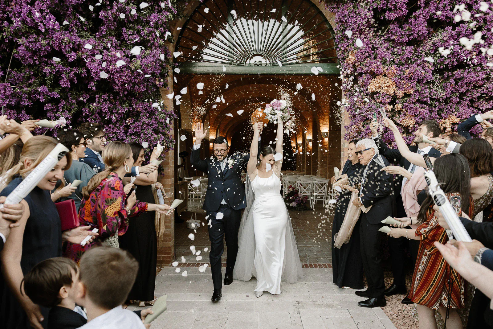 Amidst lavender-hued floral walls in Puglia, a just-married couple is showered with petals by joyful guests outdoors.