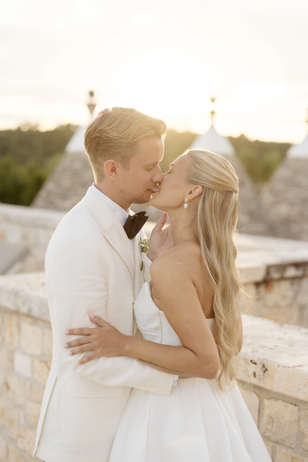 Newlyweds share a sunset kiss by ancient stone trulli in Puglia, Italy, capturing romance and timeless elegance.