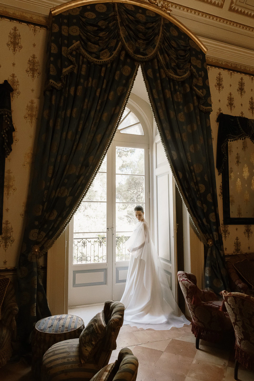In an exquisite Puglia villa, a bride in a flowing white gown gazes out a grand window draped with ornate curtains.