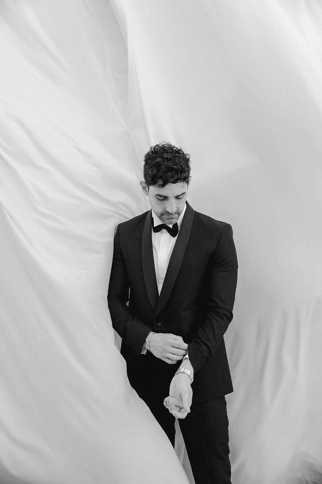 A groom in a sleek tuxedo adjusts his cuff, framed by luminous drapes—timeless elegance for a Puglia wedding editorial.