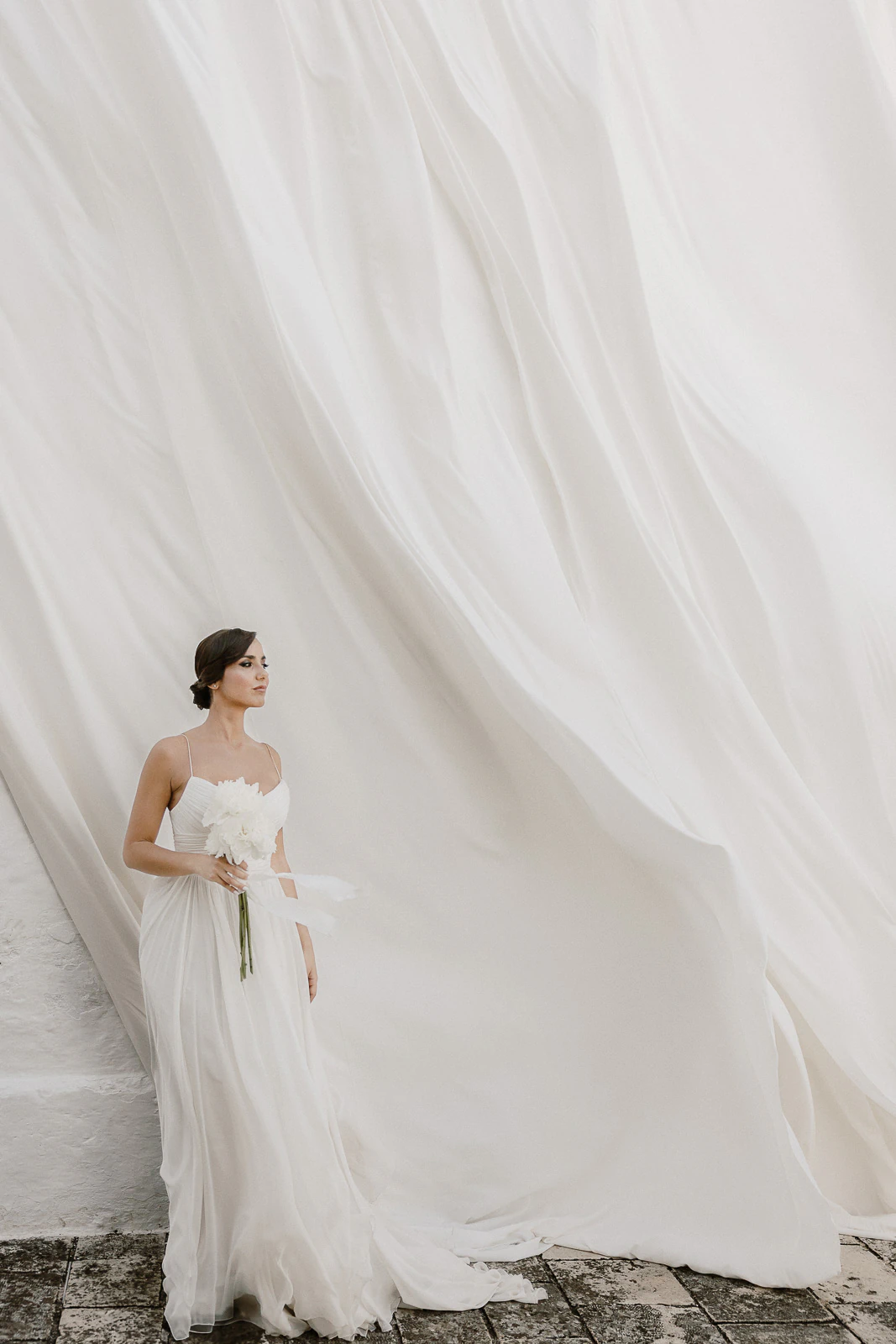 A bride in an elegant white gown holds fresh blooms, framed by flowing fabric in the soft light of a Puglia wedding.
