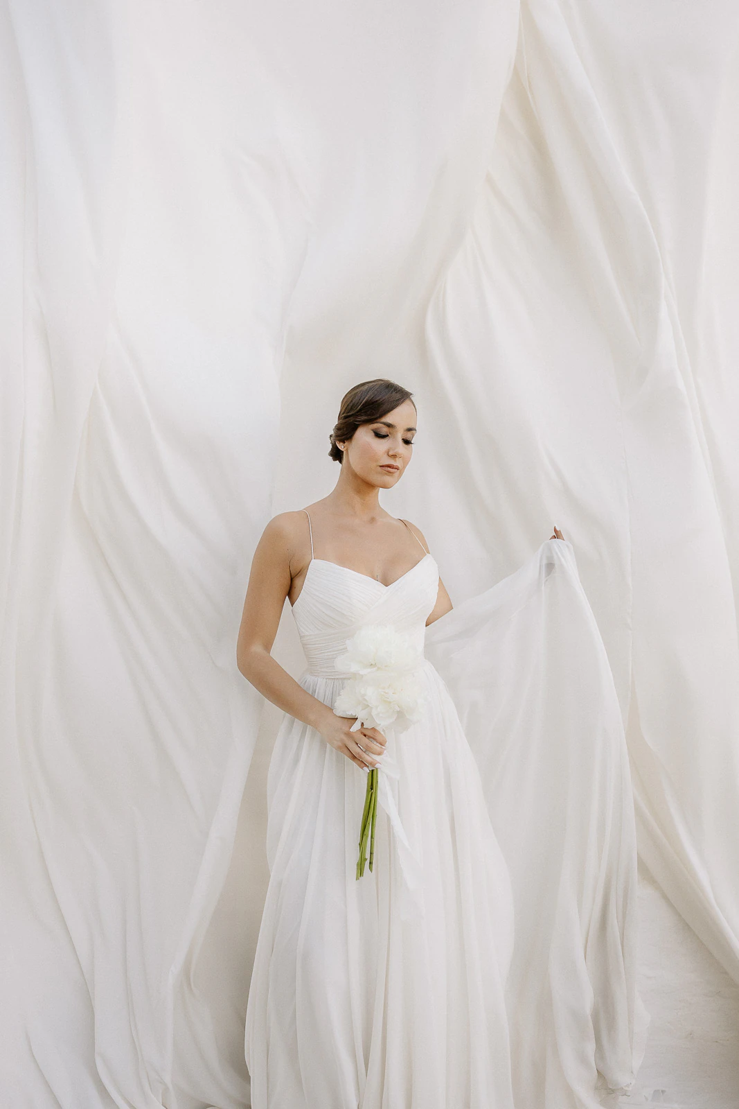 A bride in an elegant white gown gently holds a delicate bloom, framed by ethereal drapery in sunlit Puglia, Italy.