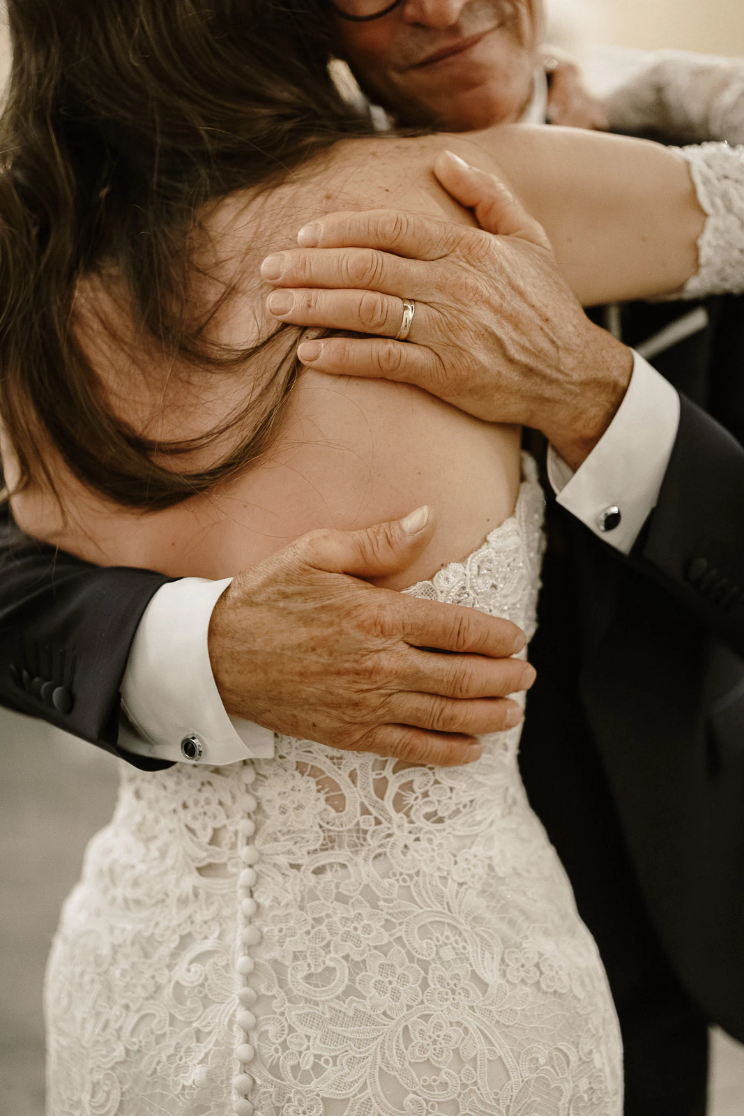 An intimate embrace between a bride in lace and her father, their hands intertwined amidst a sunlit celebration in Puglia, Italy.