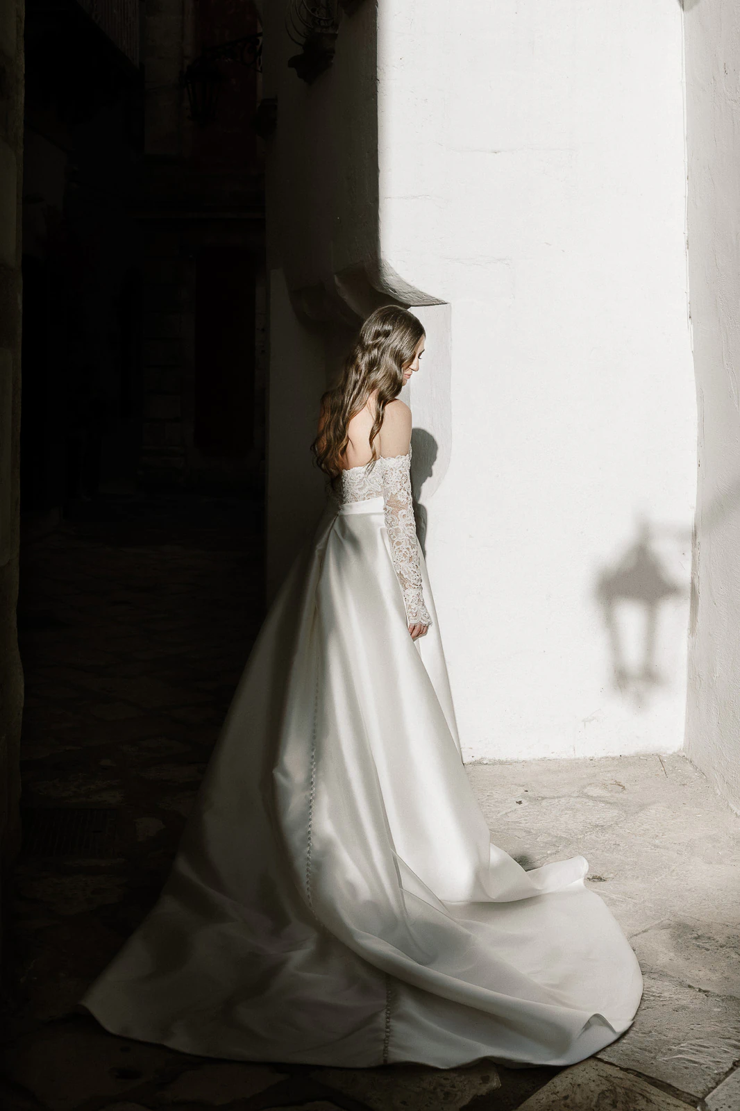 A bride in a graceful, long-sleeved gown pauses in a sunlit Puglian alley, embracing the romance of Italy.