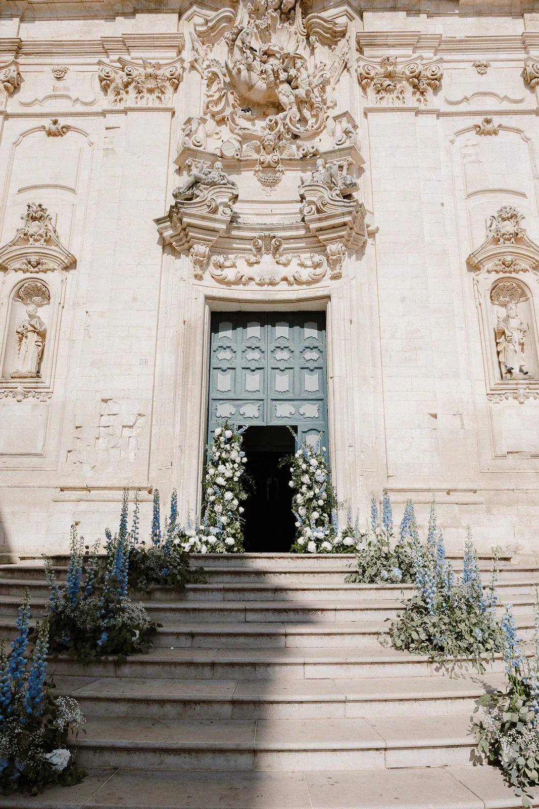 Elegant stone church in Puglia with a grand teal door, lush florals, and blue blooms framing the wedding entrance.