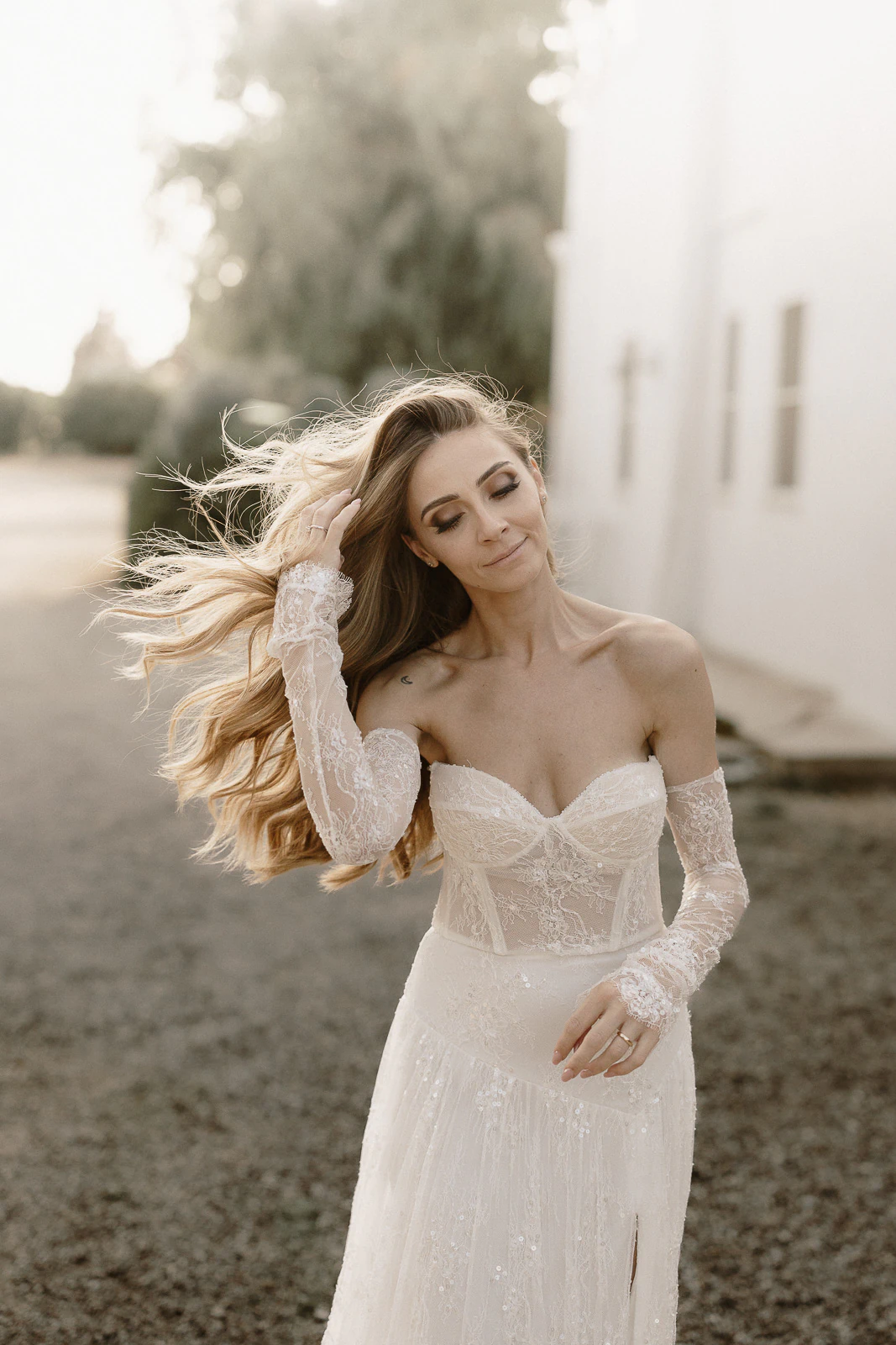 A bride in an elegant off-shoulder gown gently touches her hair amid the sunlit romance of a Puglian countryside wedding.