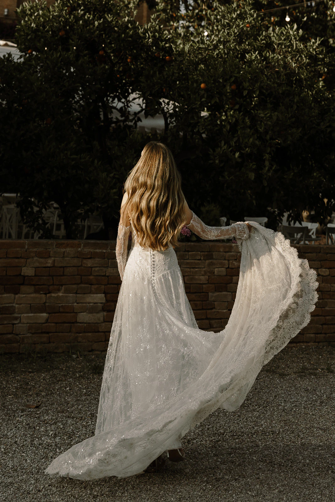 A bride in an elegant lace gown lifts her train amid sunlit Italian gardens and rustic stone, capturing destination romance.