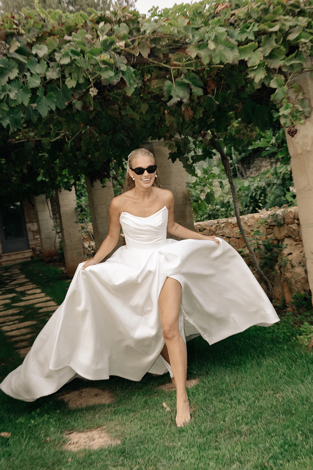 A bride in sunglasses strolls barefoot beneath a vine-draped pergola in Puglia, radiating joy and effortless Mediterranean elegance.