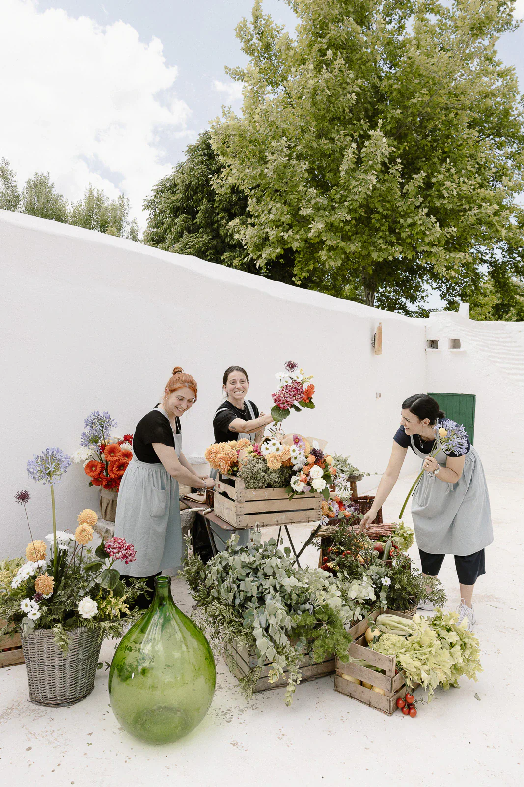 In sunlit Puglia, three women artfully arrange blooms amid lush greenery and whitewashed walls, capturing Italian wedding elegance.