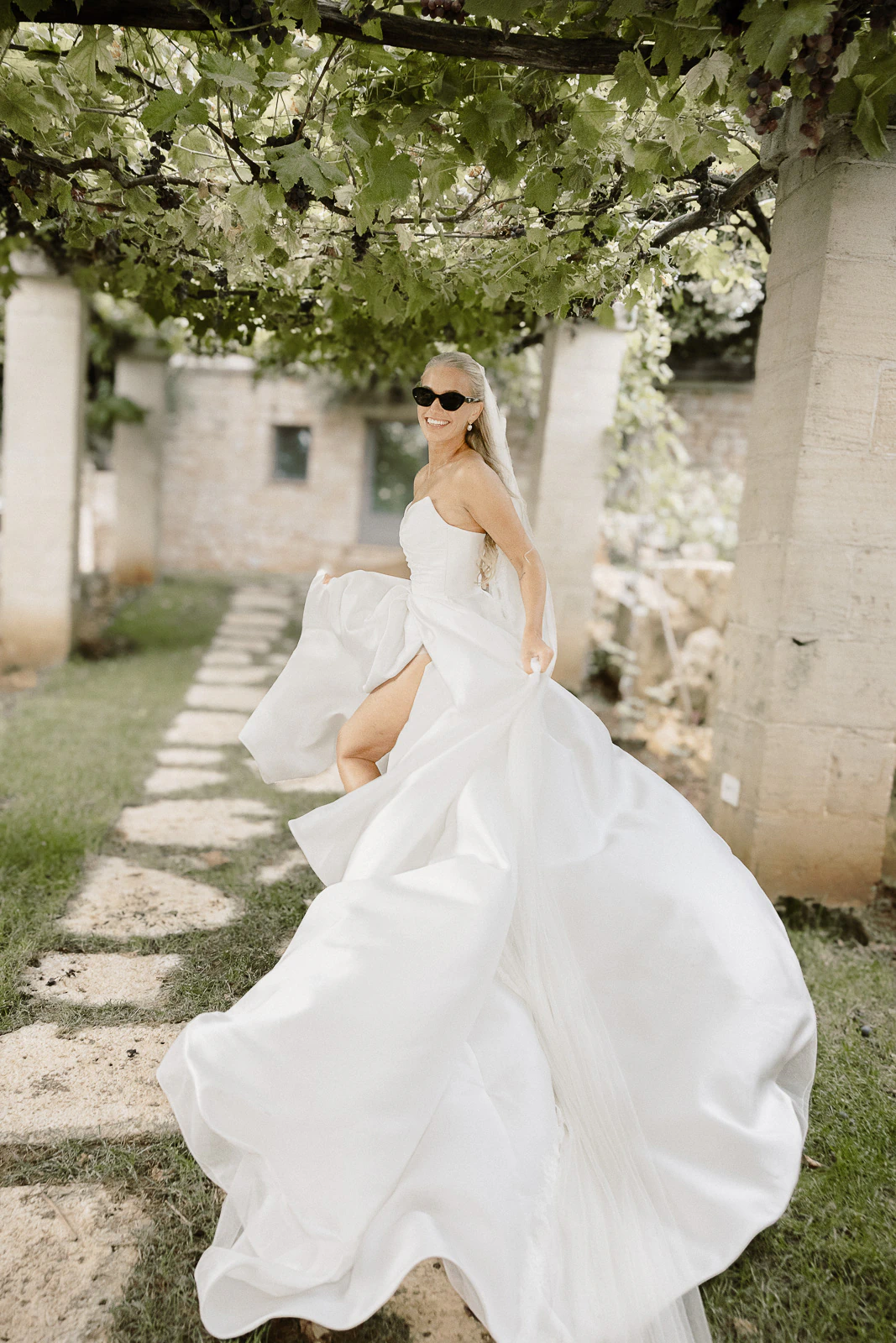 A bride in sunglasses joyfully twirls beneath a leafy pergola and stone columns, embracing the sunlit romance of Puglia, Italy.