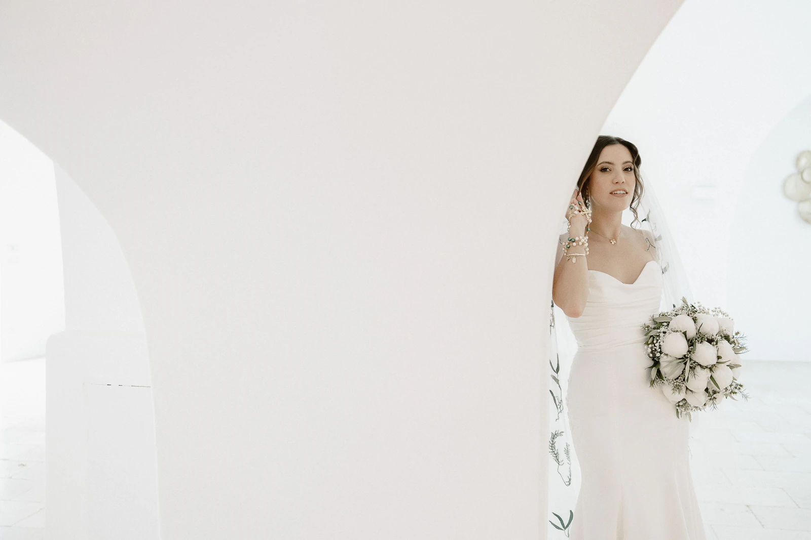 A bride in an elegant white gown stands serenely by a sculptural arch, bathed in soft Apulian light.