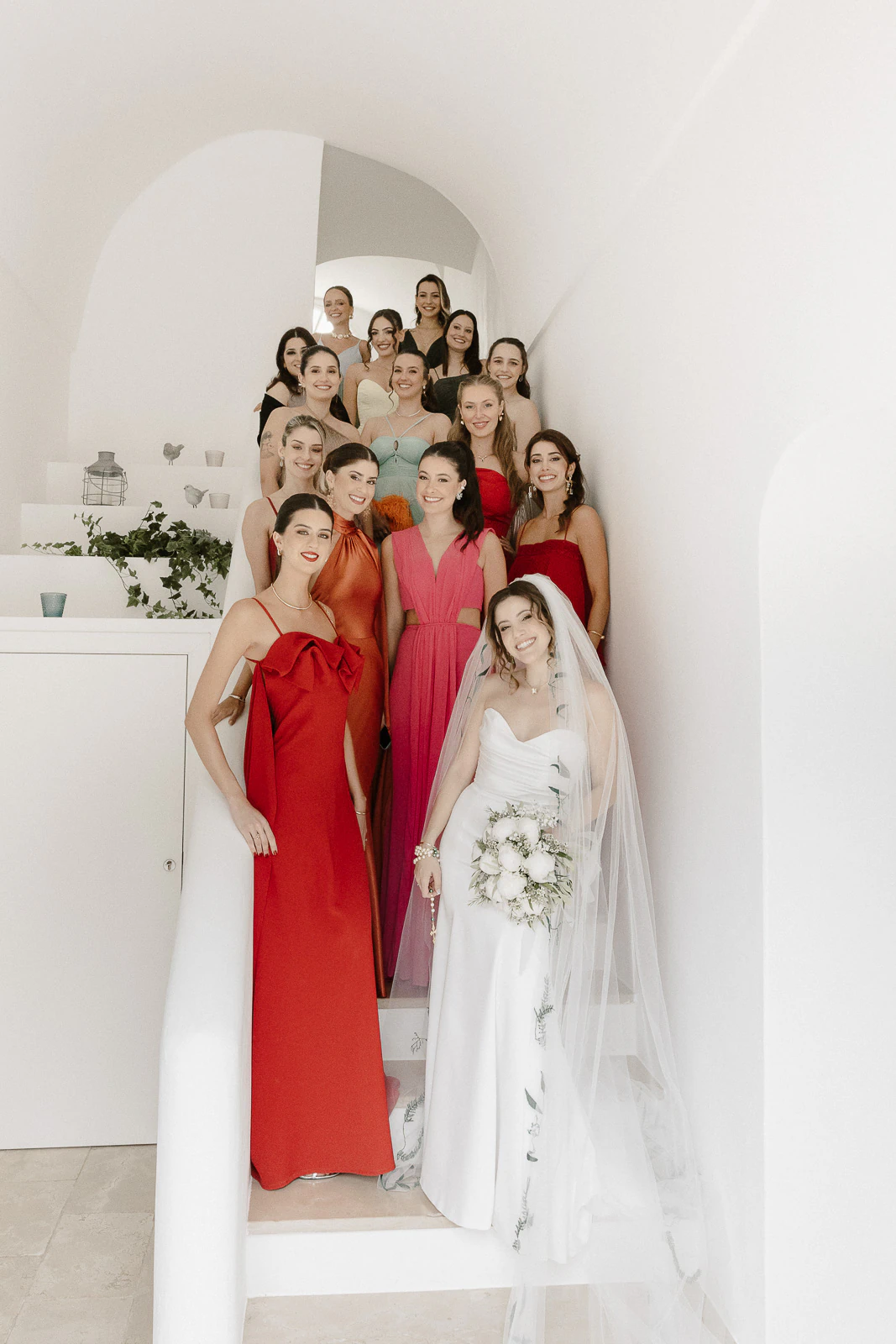 In sunlit Puglia, a radiant bride ascends minimalist white stairs, her bridesmaids in vibrant hues gathering joyfully behind her.
