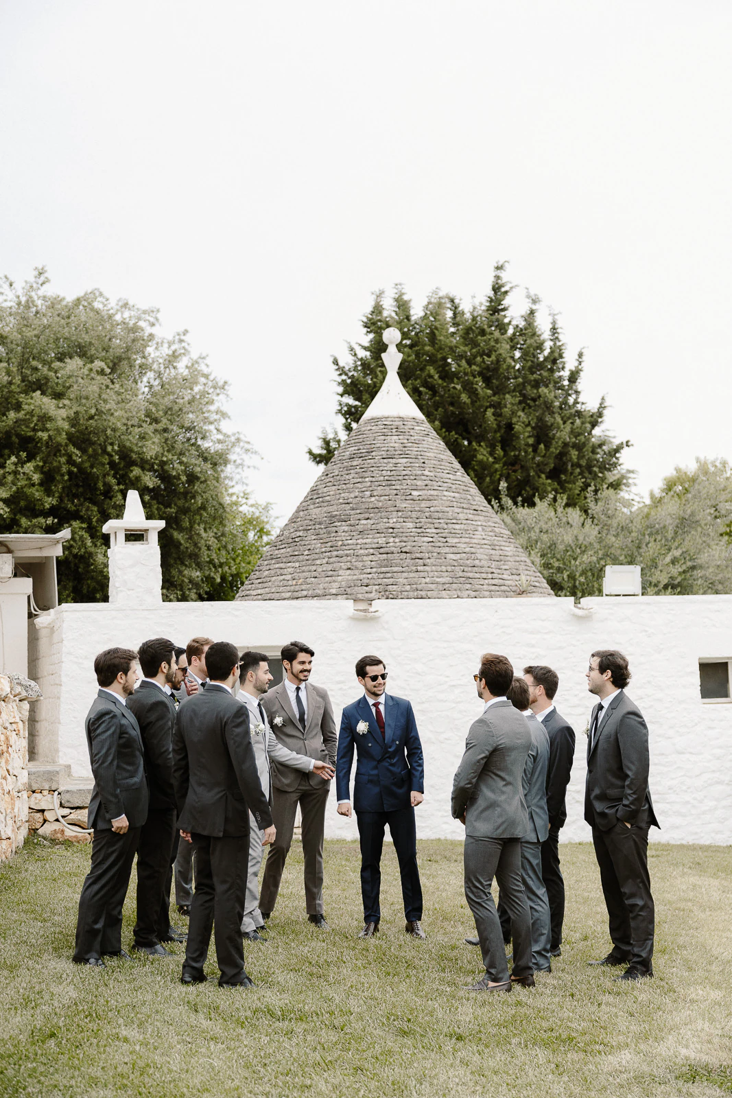Groomsmen share a candid moment on lush lawns before a white trullo, embracing the charm of a Puglia wedding setting.