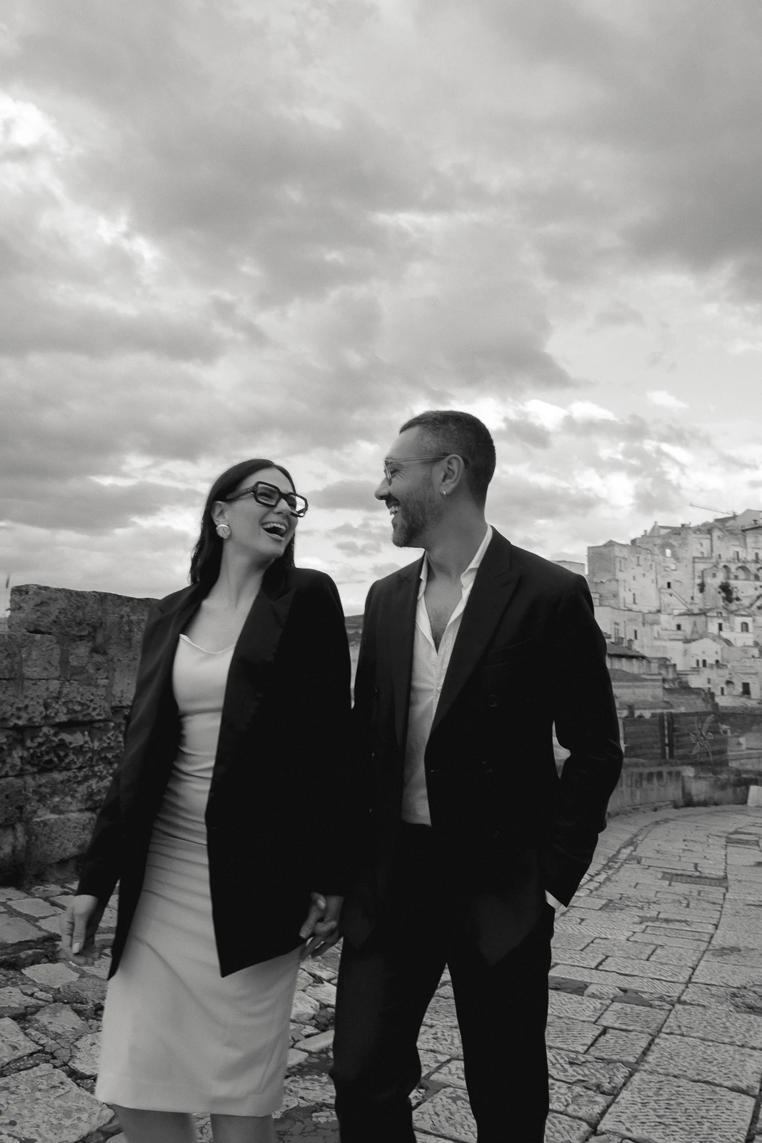 An elegantly dressed couple strolls a stone path in Puglia, Italy, sharing joyful smiles beneath a romantic, overcast sky.