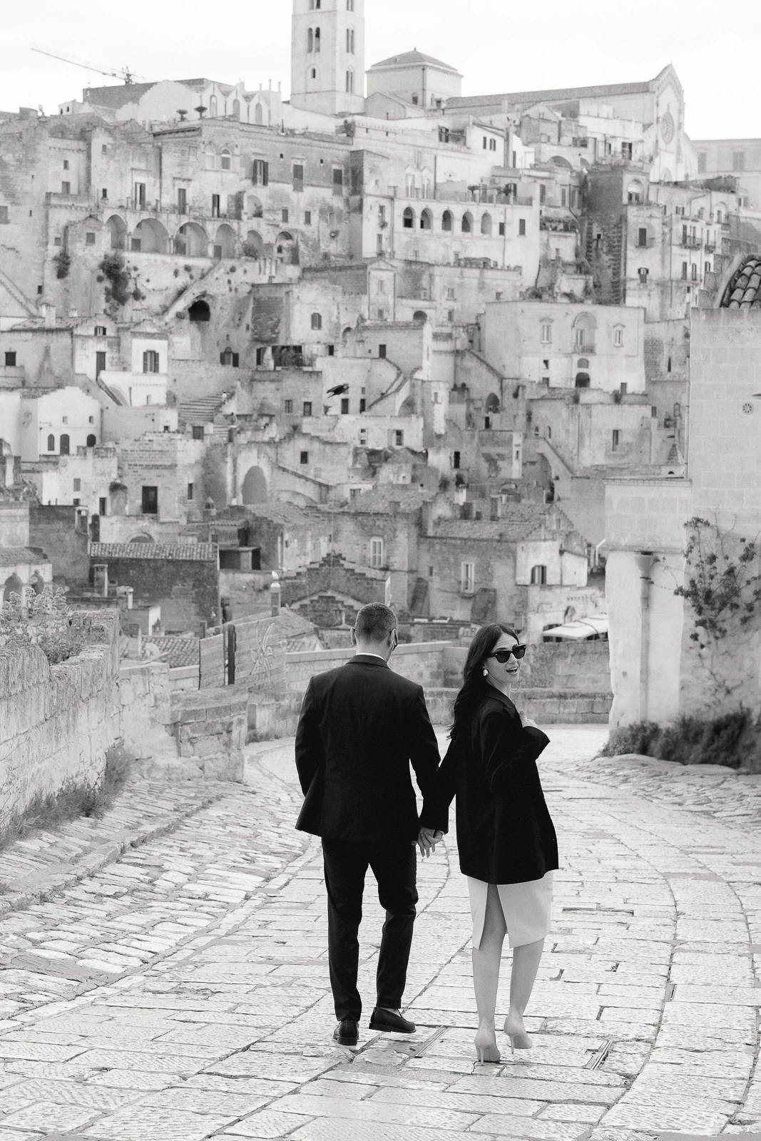 Elegantly dressed couple walk hand in hand along a sunlit stone street, historic Puglia hillside buildings rising behind them.