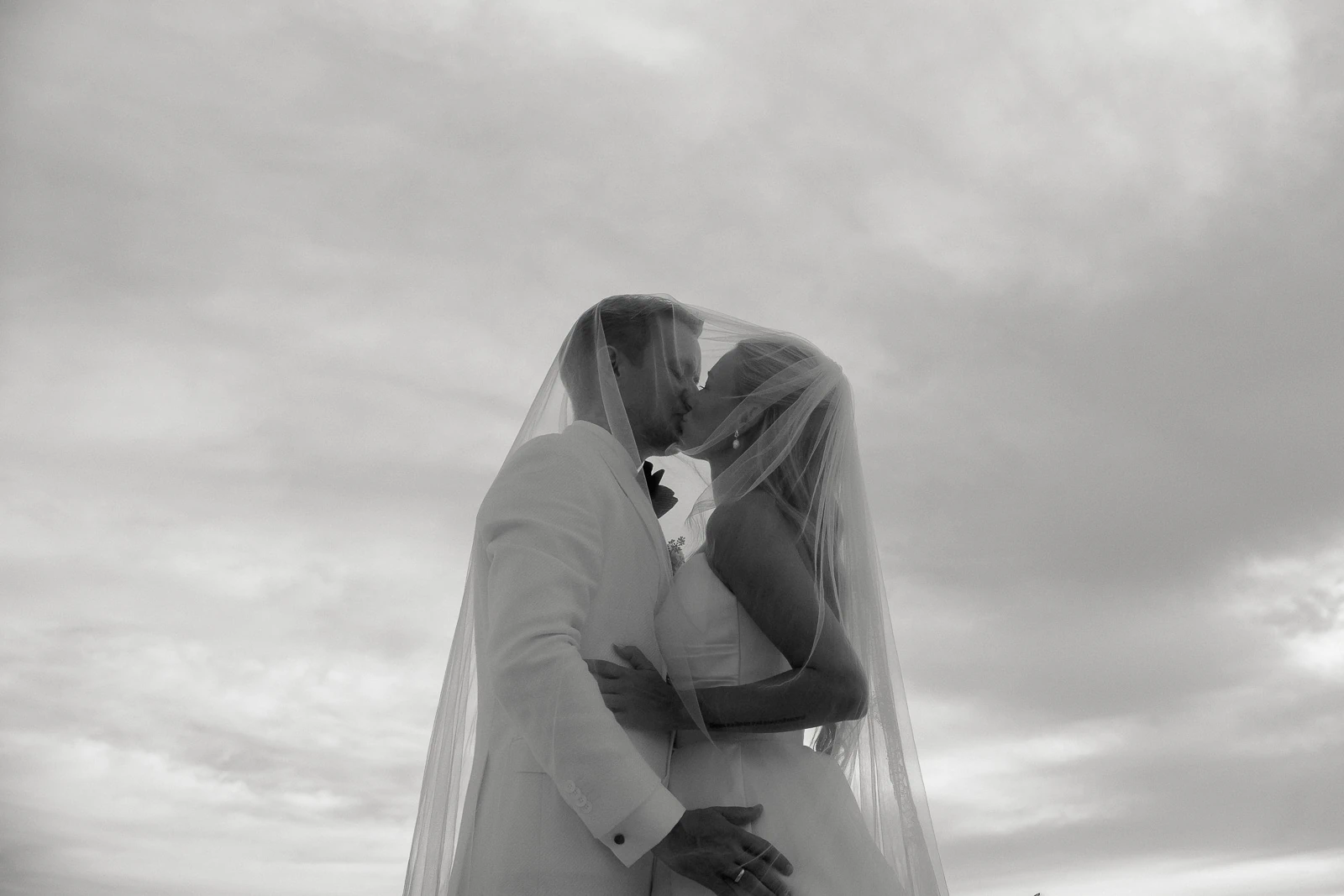 Beneath soft Puglian skies, a bride and groom share an intimate kiss under a veil, wrapped in timeless elegance.