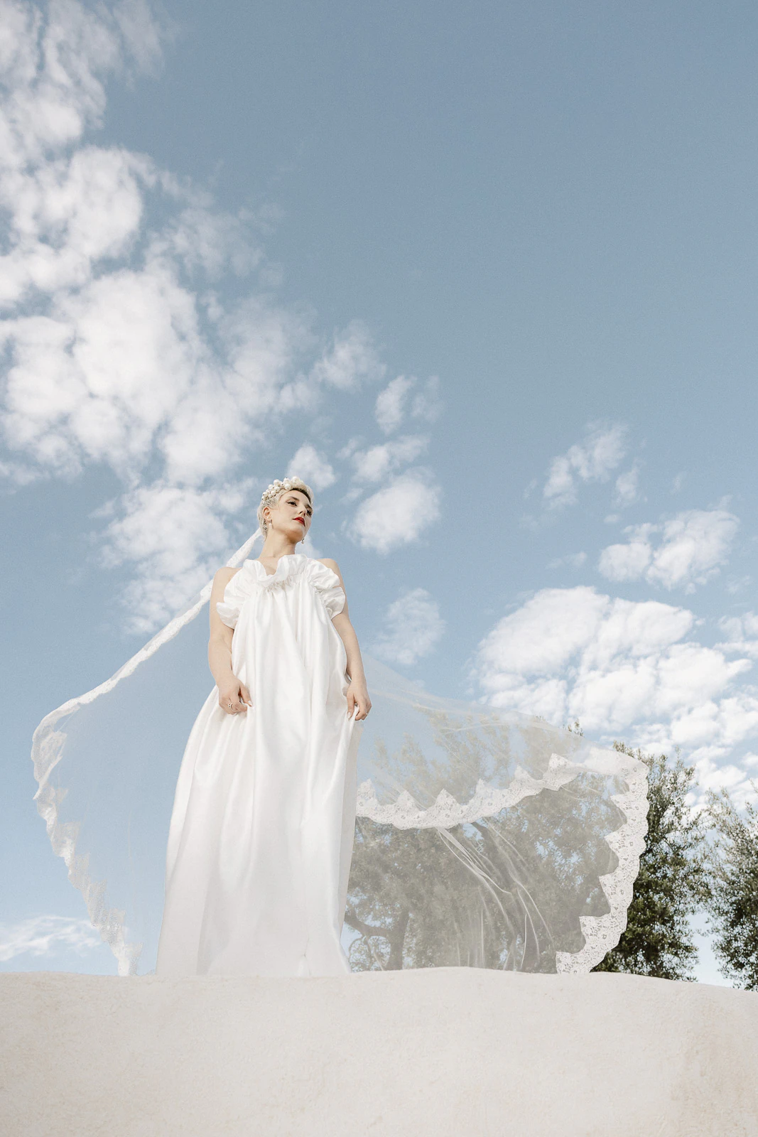 Beneath the open Puglian sky, a bride in delicate lace gazes thoughtfully, embraced by Italy’s serene countryside elegance.