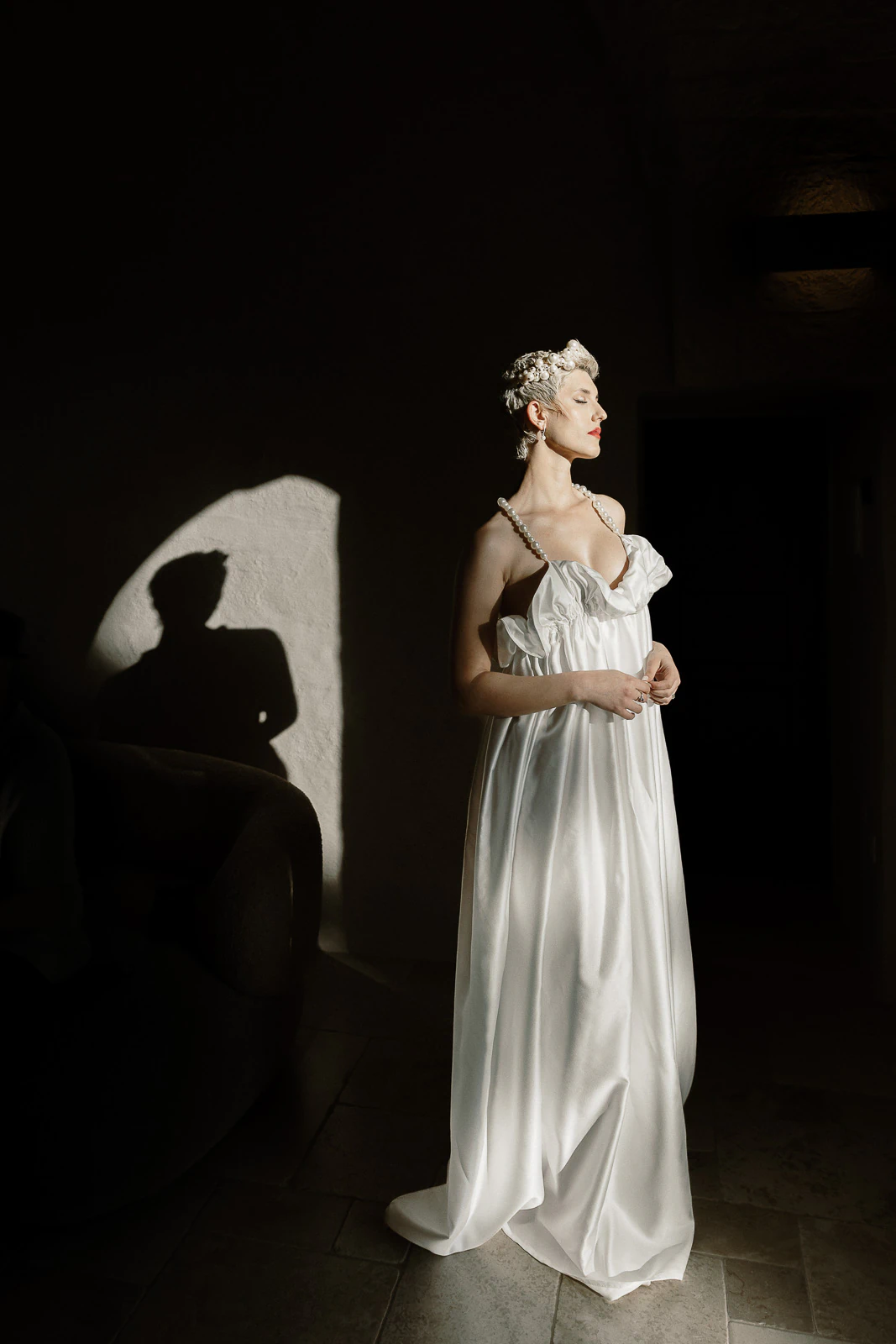Bathed in ethereal light, a bride in white stands serenely in a dim Apulian room, her shadow dancing on ancient walls.