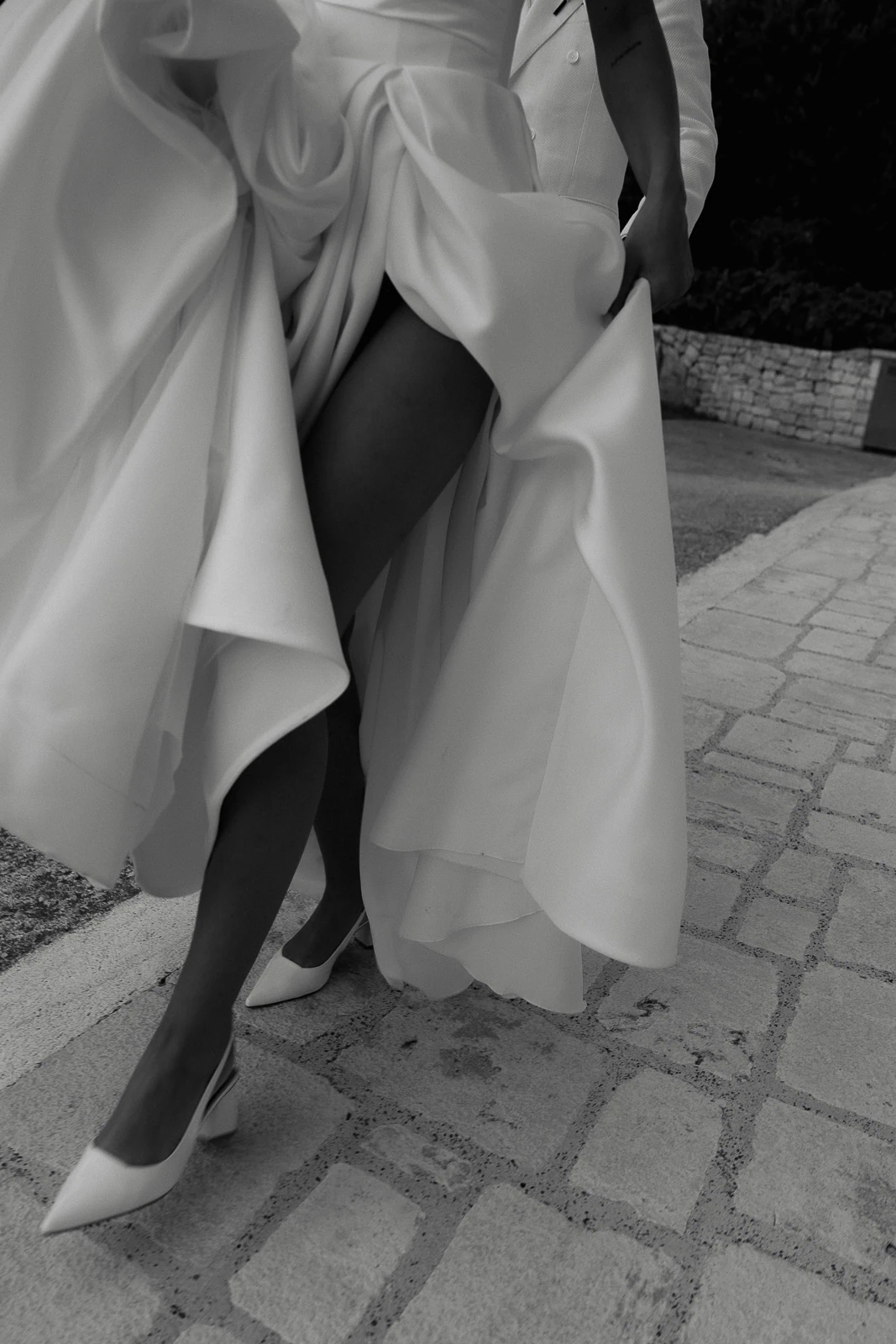 A bride in a flowing white dress strolls a sunlit stone path in Puglia, Italy, hand-in-hand with her partner.