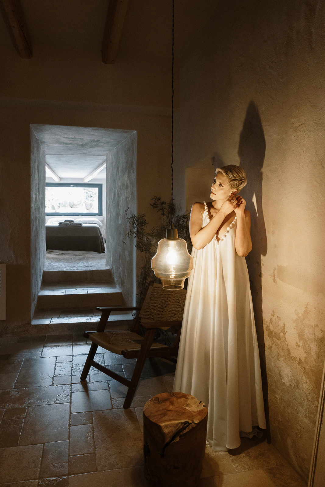 A bride in a flowing white gown adjusts her earring beside a glowing lamp in an intimate Puglia stone villa.